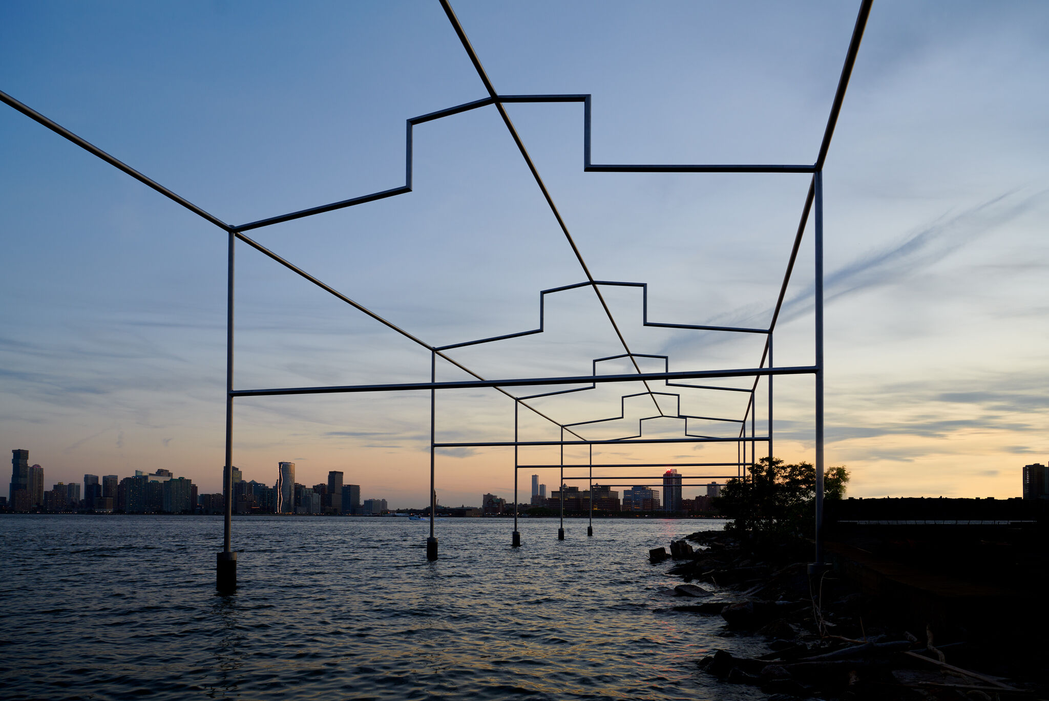 A large stainless steel structure—the outline of a pier—hovers over the Hudson River at dusk. 