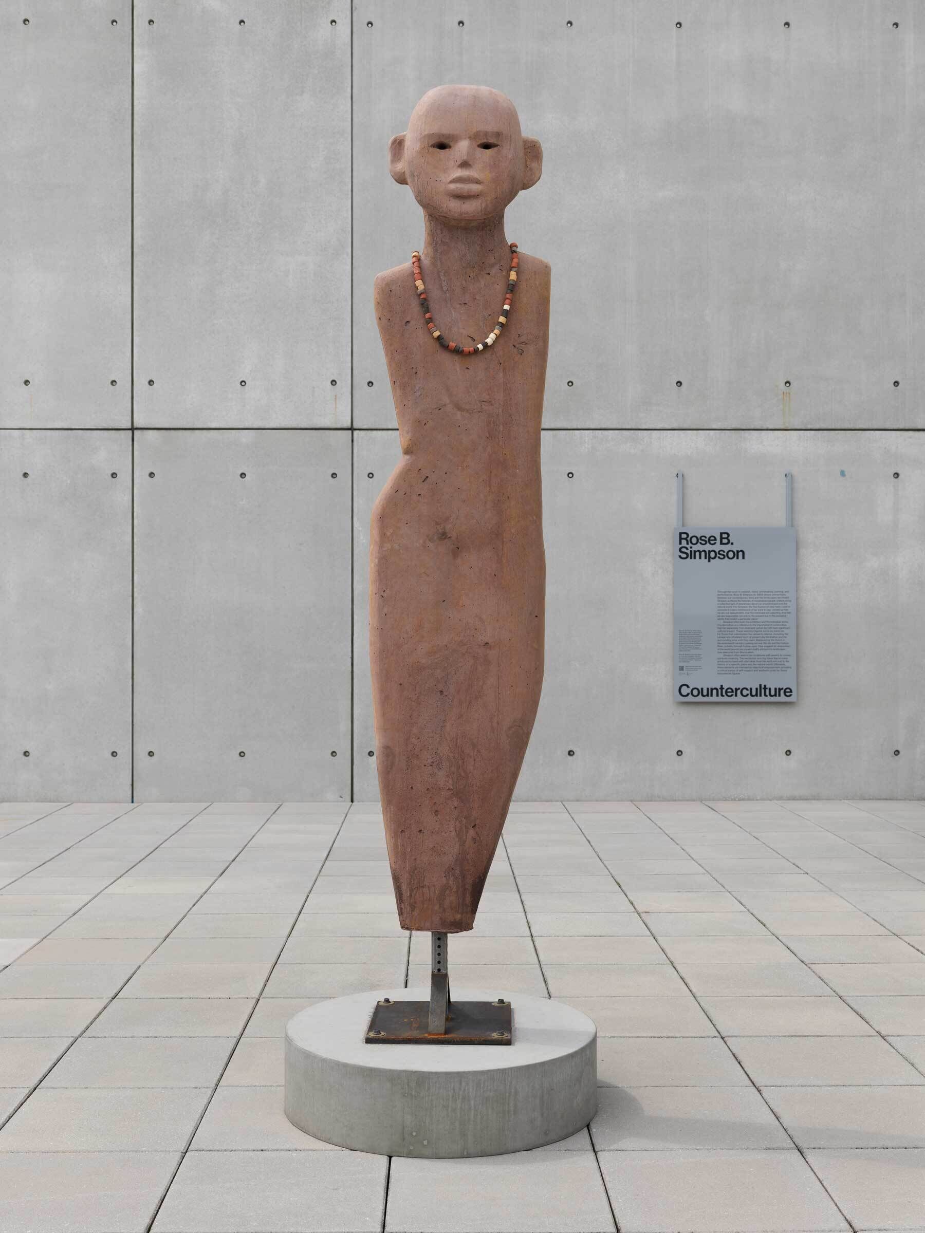 A tall, brown human figure sculpture wearing beaded necklaces with no limbs stand straight on the balcony.