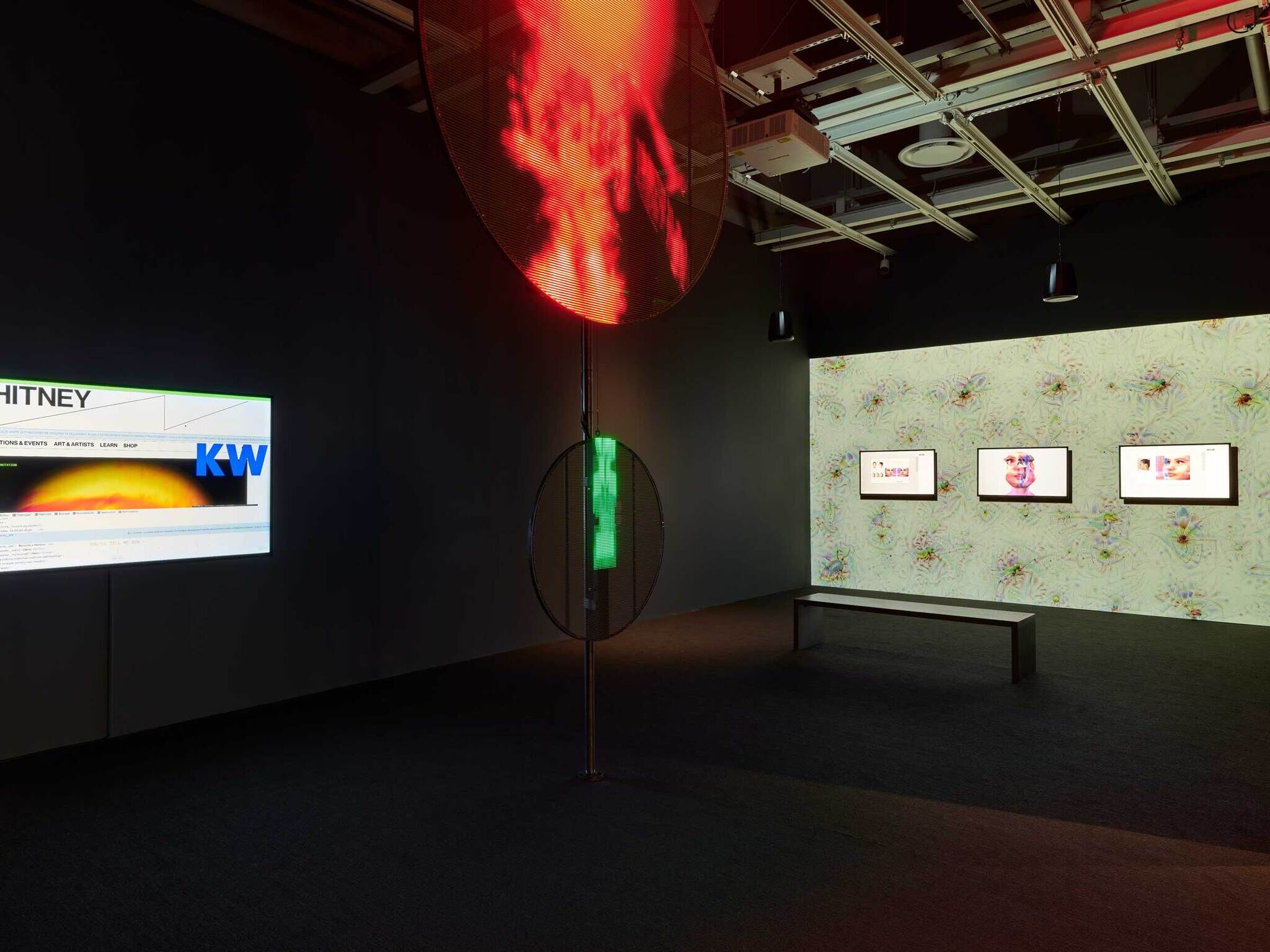 A dark room with a screen on the left to a Whitney museum page, two dark circular LED lights one red and one green, and a big screen with three smaller screens on it on the right with a green background and humanoid faces on the smaller screens.