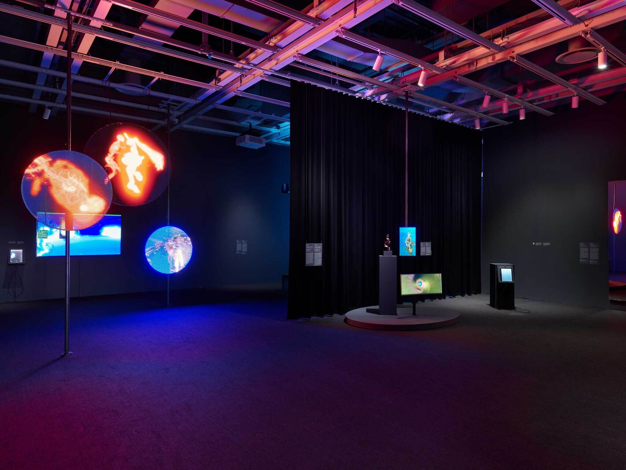 A gallery view of the exhibition with three circular glowing LED screens. To the right, there is a small sculpture next to two computer screens, and a big box machines with a screen all in front of a curtain with a light coming out behind it.