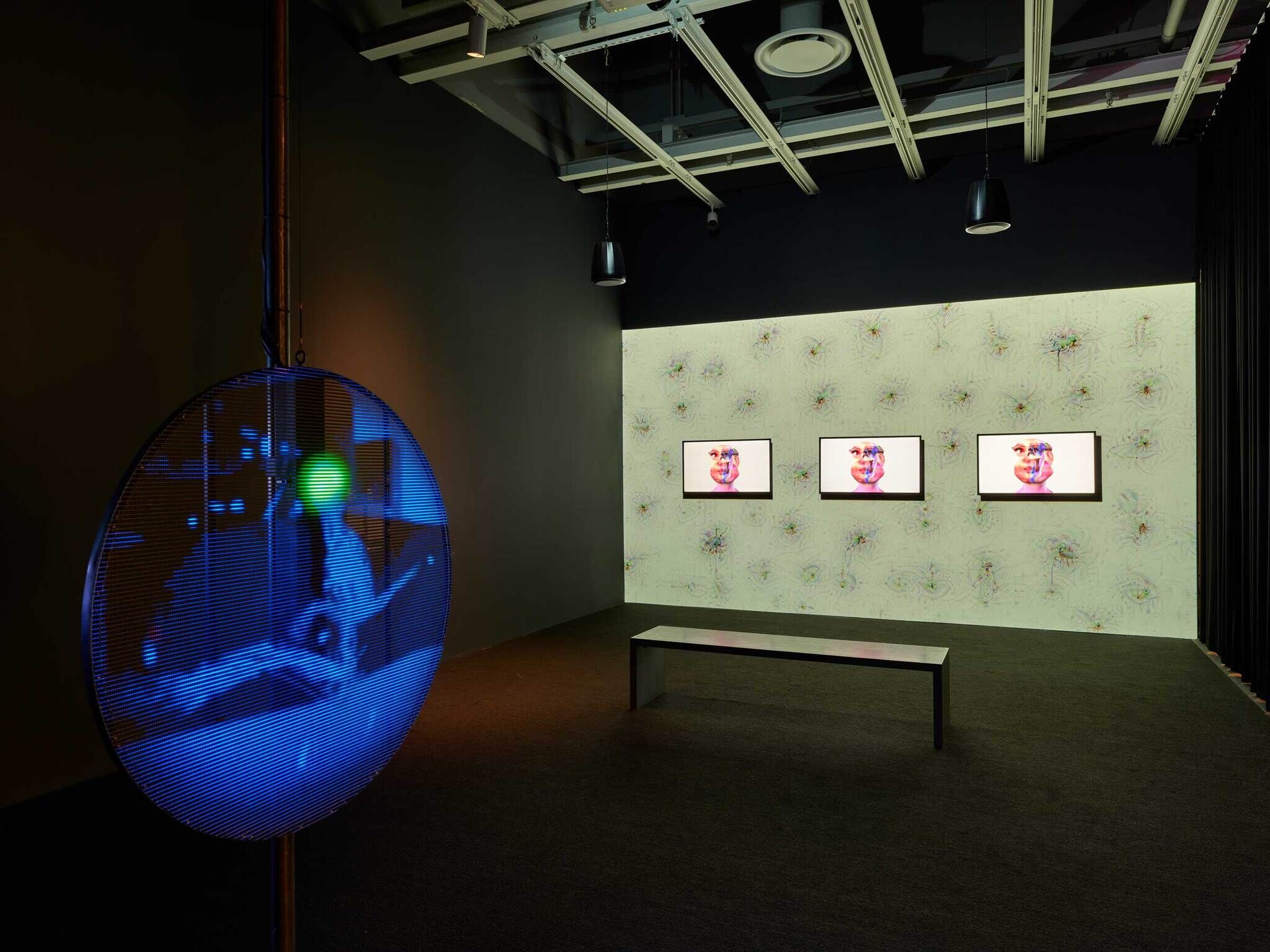 A blue glowing circle in the foreground and in the background the big screen with a green digital pattern behind three smaller screens with humanoid faces.