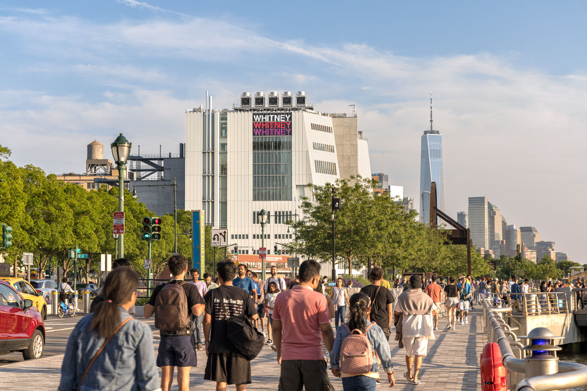 A view of the Whitney Museum from the walkway along the Hudson River by the West Side Highway in New York. There are crowds of people walking south along the path and the Whitney is visible on the left side and the Freedom Tower in the distance on a sunny day with blue skies. 