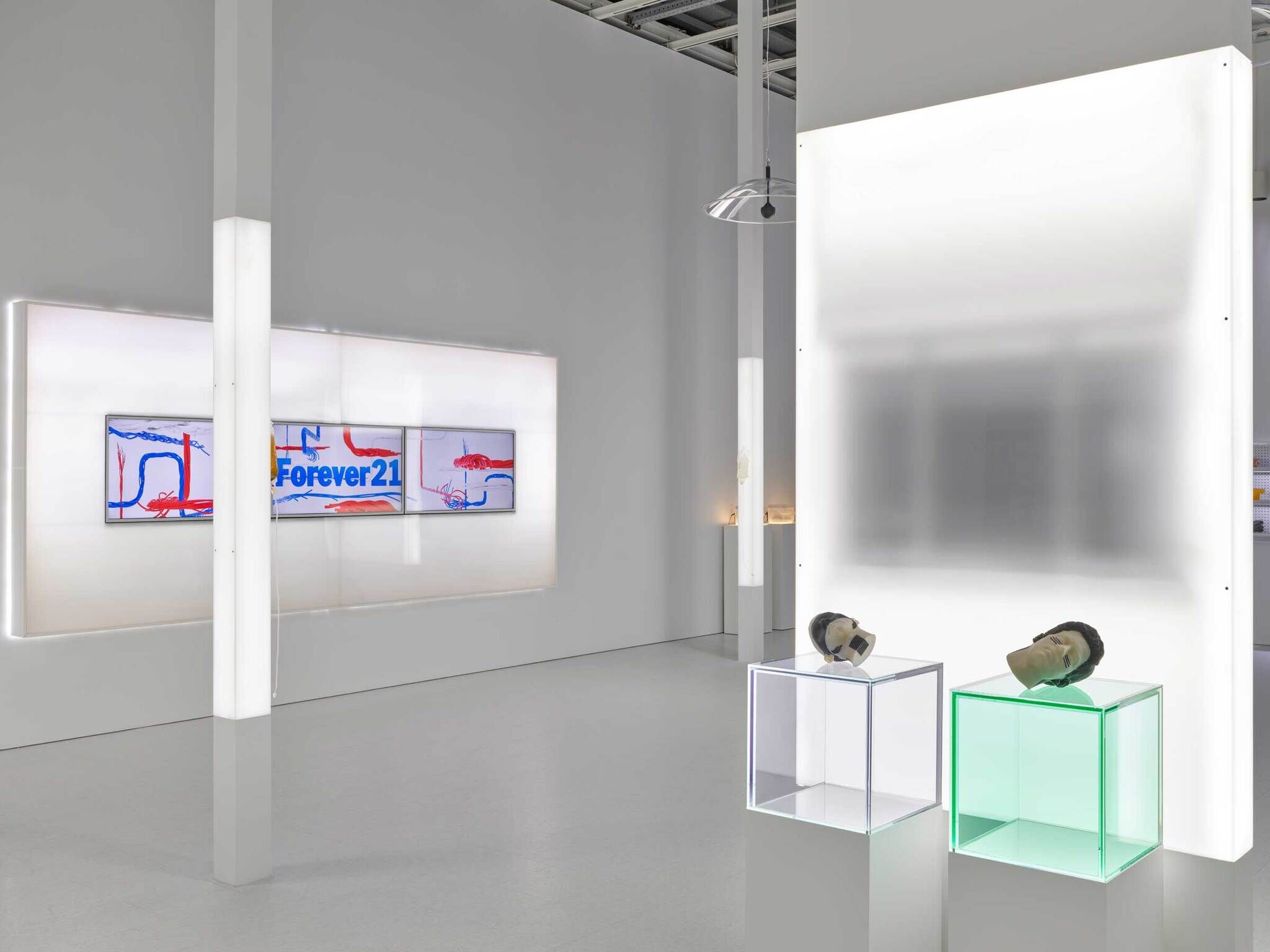 A grey room with light panels, on the left there is a triptych of red and blue designs with the words Forever21, and on the right there are two transparent boxes with mannequin heads.