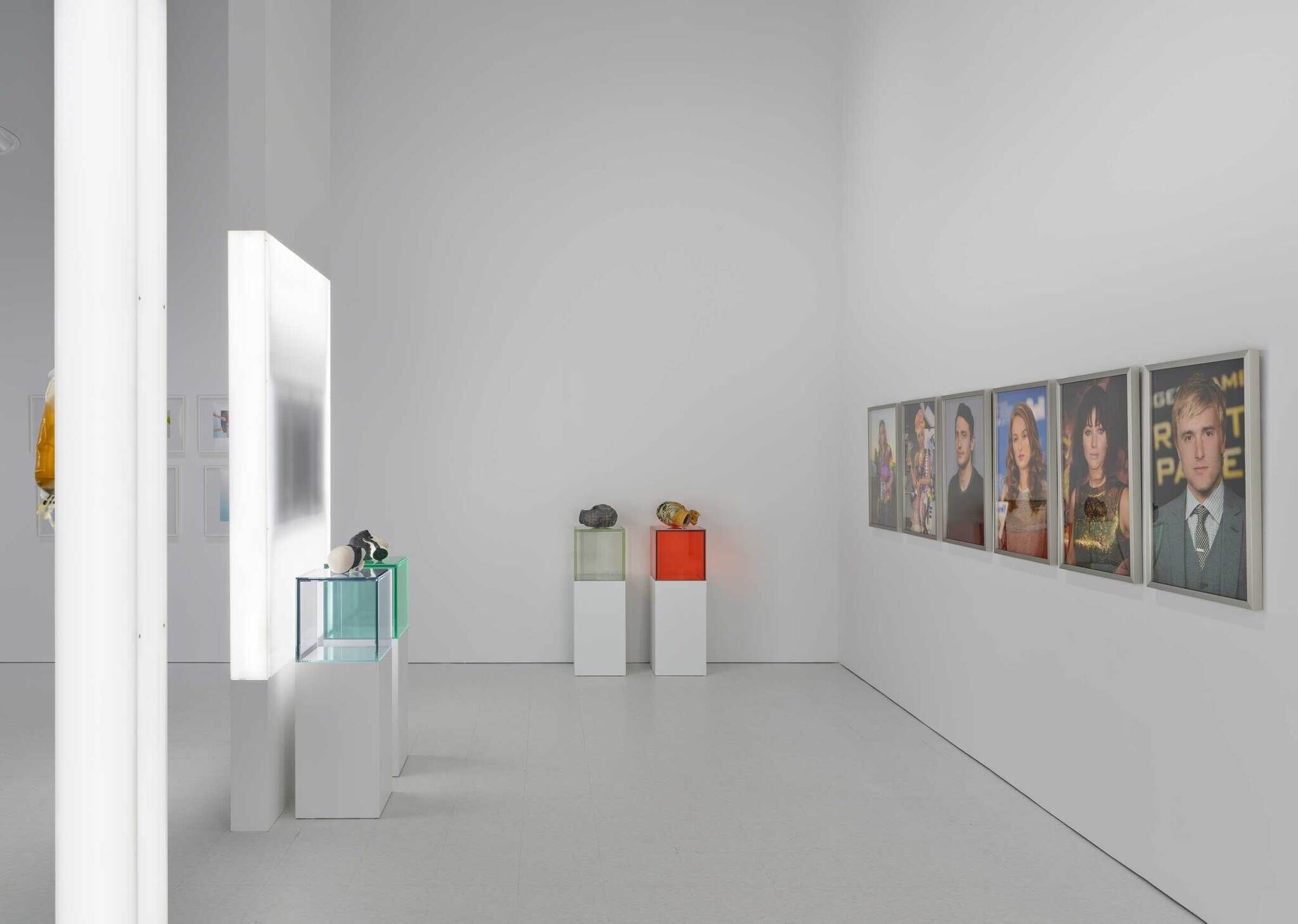 From left to right: two transparent boxes with mannequin heads, two transparent boxes with mannequin heads, and six photograph portraits on the right wall.