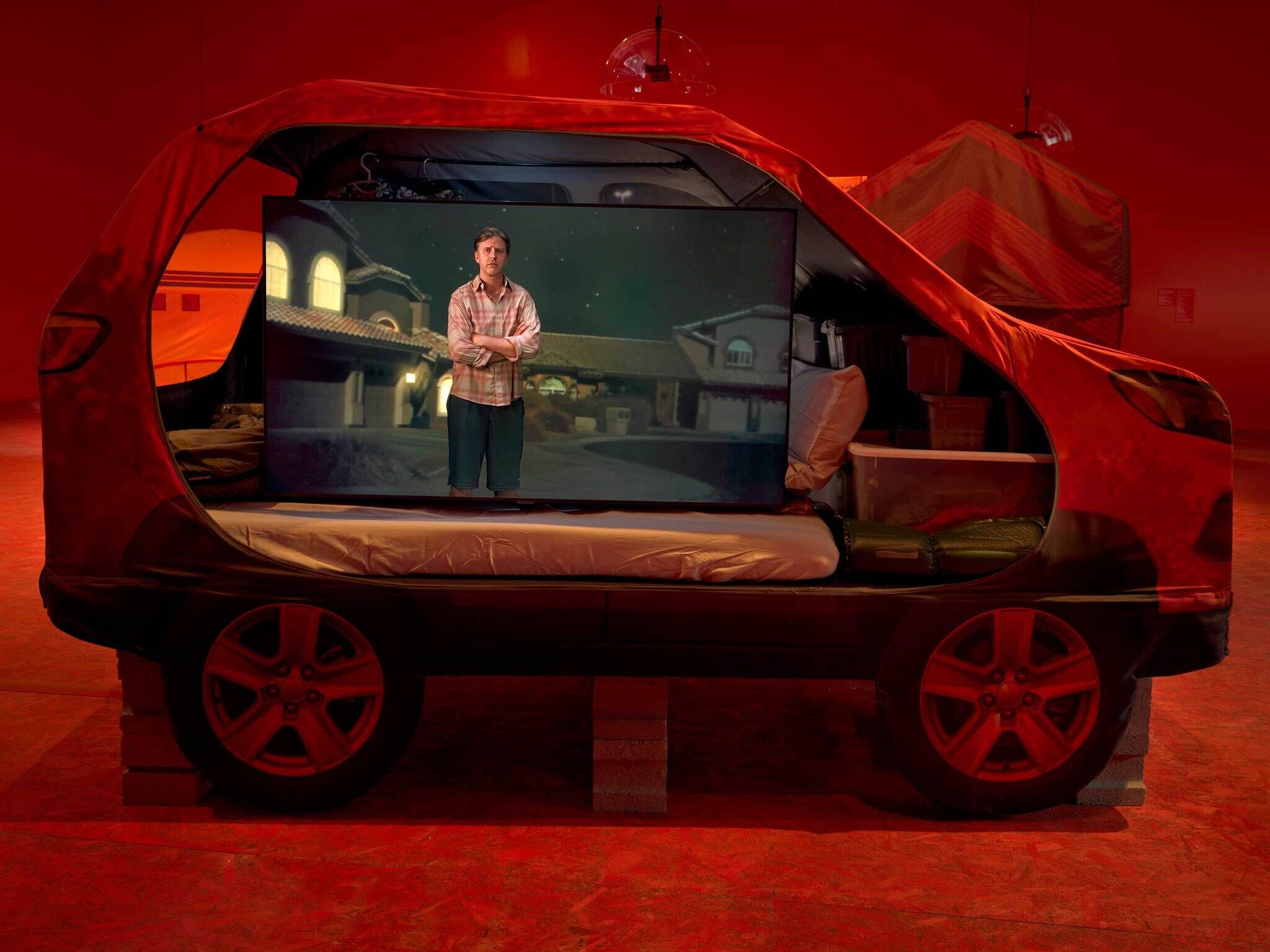 A car drenched in red light with a screen inside with an image of a man in the suburbs.