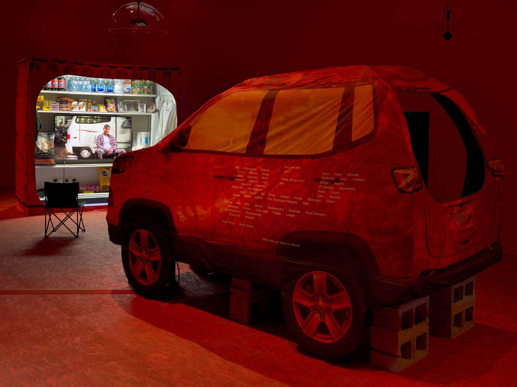 A tent car in red light on cement blocks facing a projection of a man in front of a car inside a pantry.