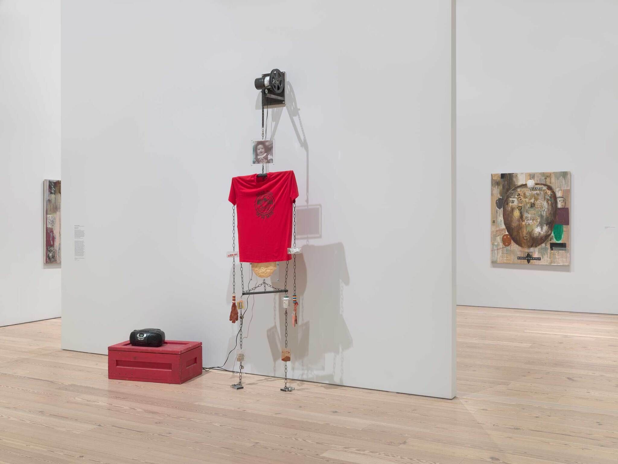 On the left there is a stereo on top of a red cargo box, on the right is a wire sculpture human figure with a red t shirt and a baby photograph as the head.