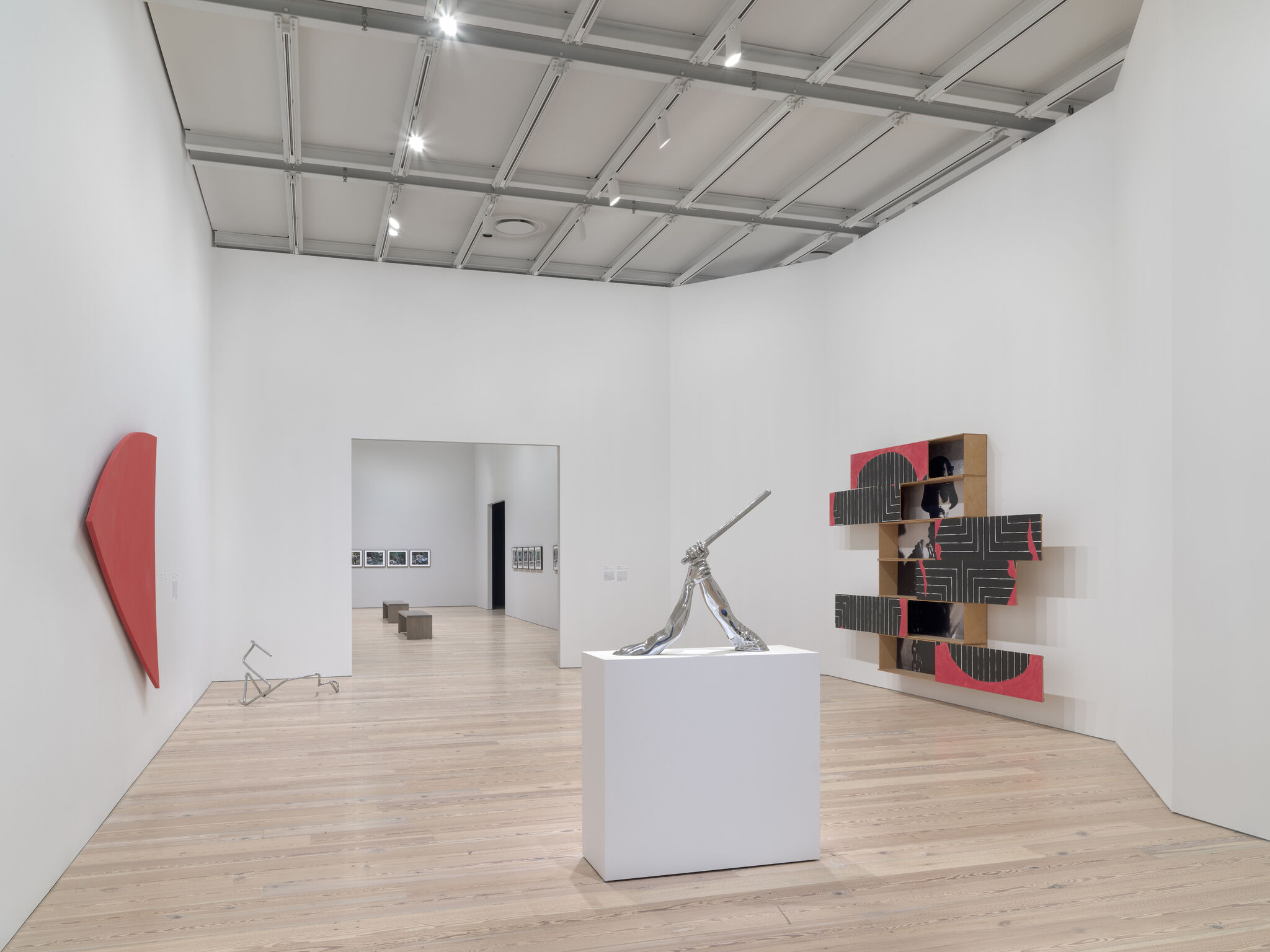 A silver sculpture on a pedestal of an arm holding a baton and another holding the wrist. On the right, a sculpture with boxes jutting out, and on the left a red abstract curved sculpture, and a wire sculpture on the ground.