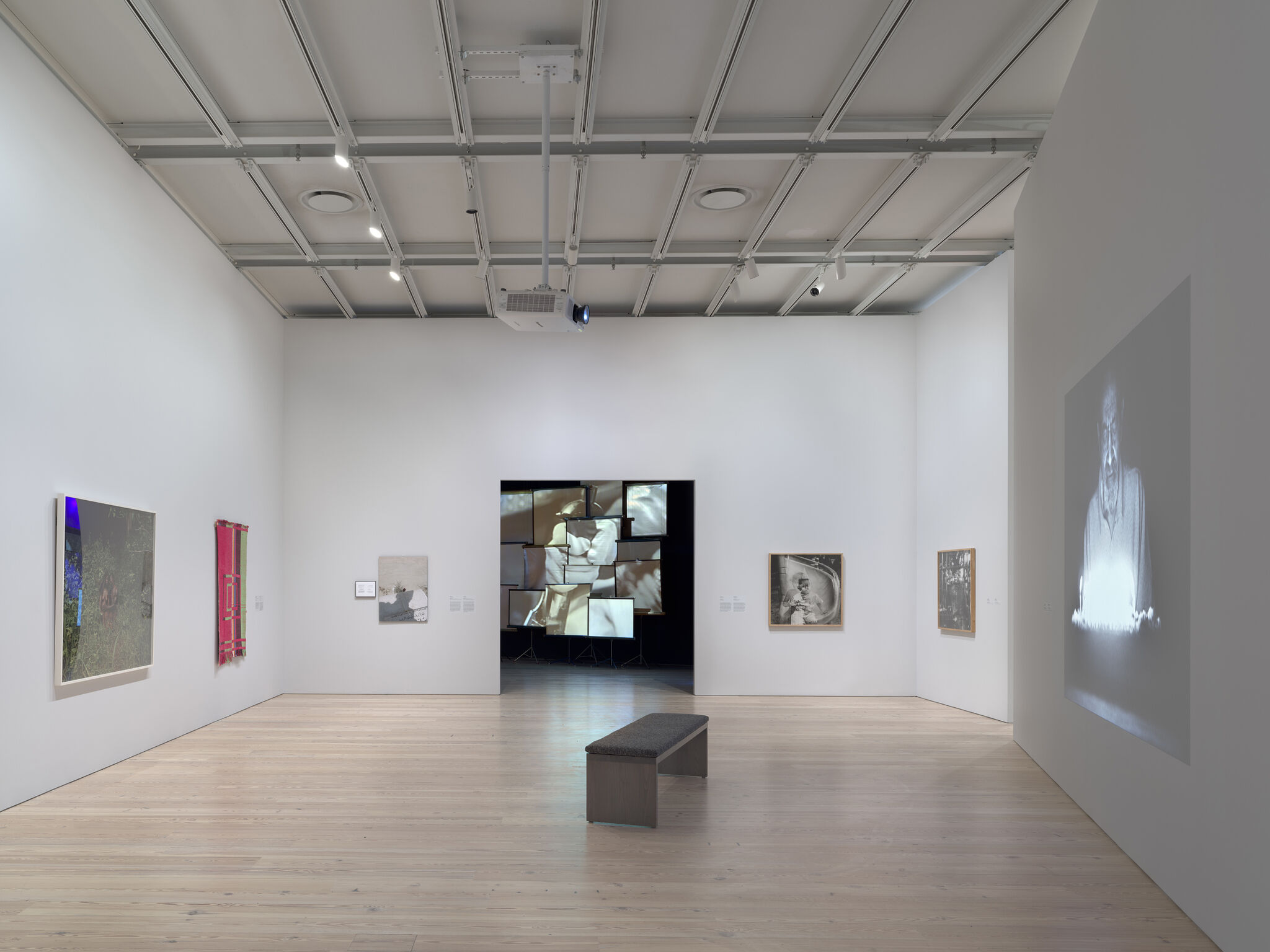 From left to right: a photograph with a blue light on it, a tapestry of pink and green, a doorway leading into a dark room with multiple screens, a black and white overlay photograph, another black and white photograph with vertical lines, and a projection of a black and white man.