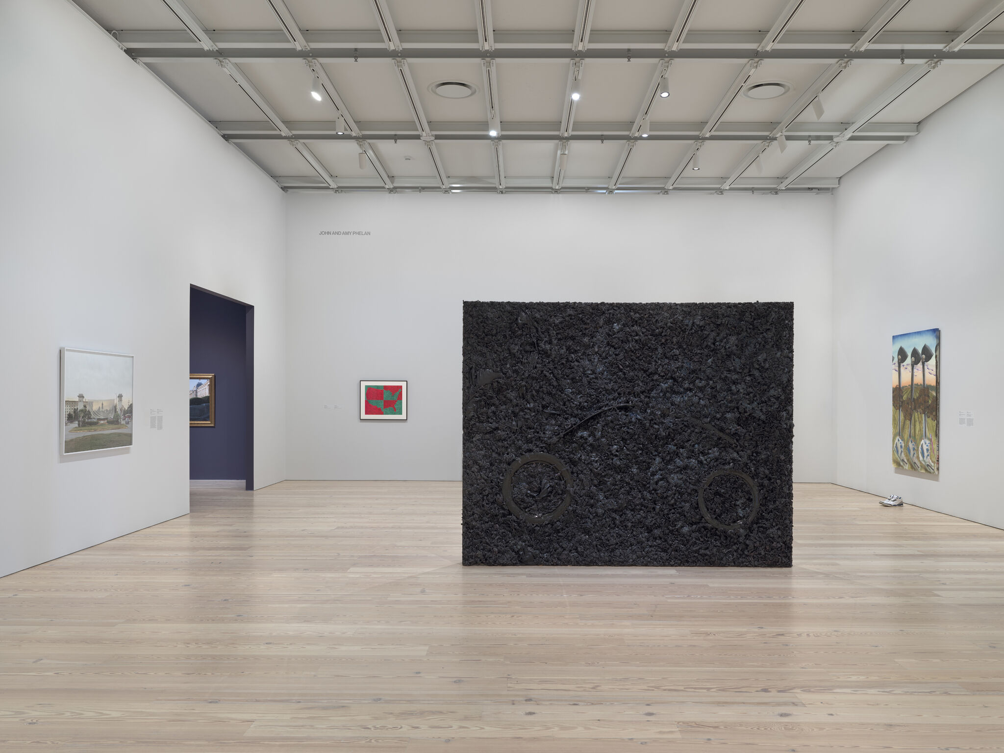 A dark textural painting in the center of the room with bike wheels. On the right, a painting with shoes placed underneath, and to the left, a red and green painting of the United States shuffled around and a landscape photograph. A hallway into another room with purple walls is in between the two.