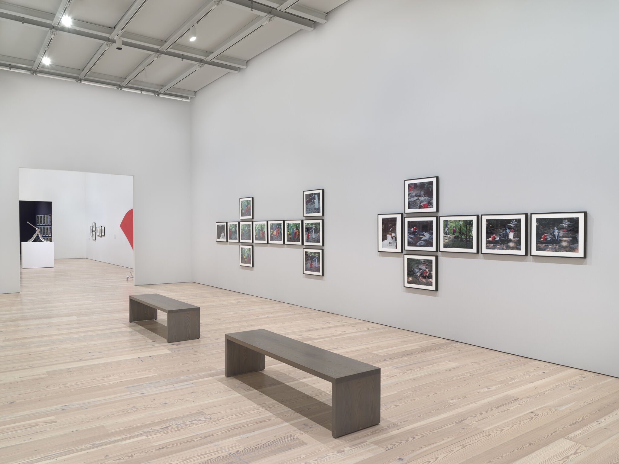 18 framed photographs placed in two sideways T-shapes with benches in front of them leading into a room with a red painting on the wall and a silver sculpture.