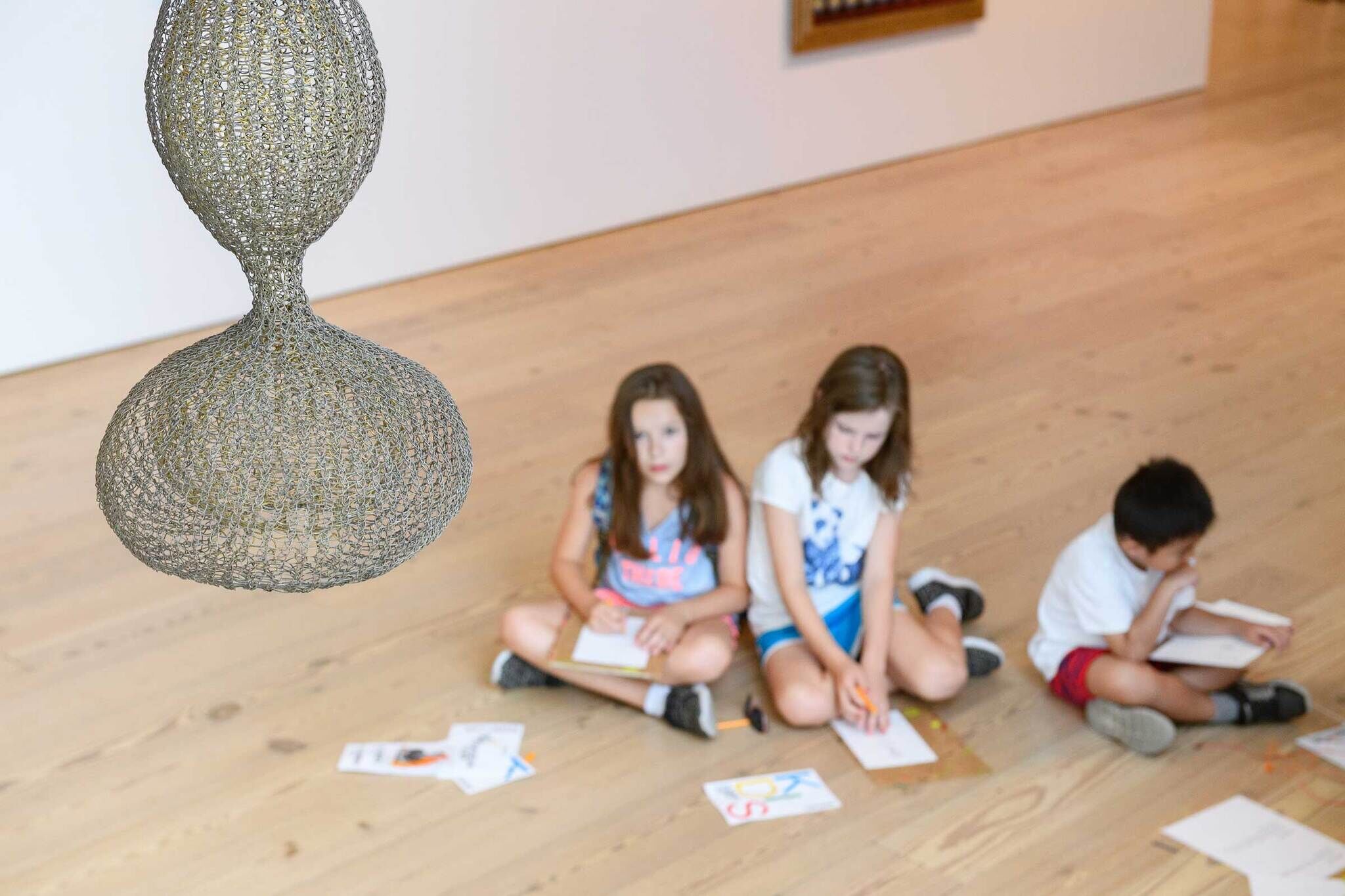 Two kids in the galleries, completing an artmaking activity before a Ruth Asawa sculpture.