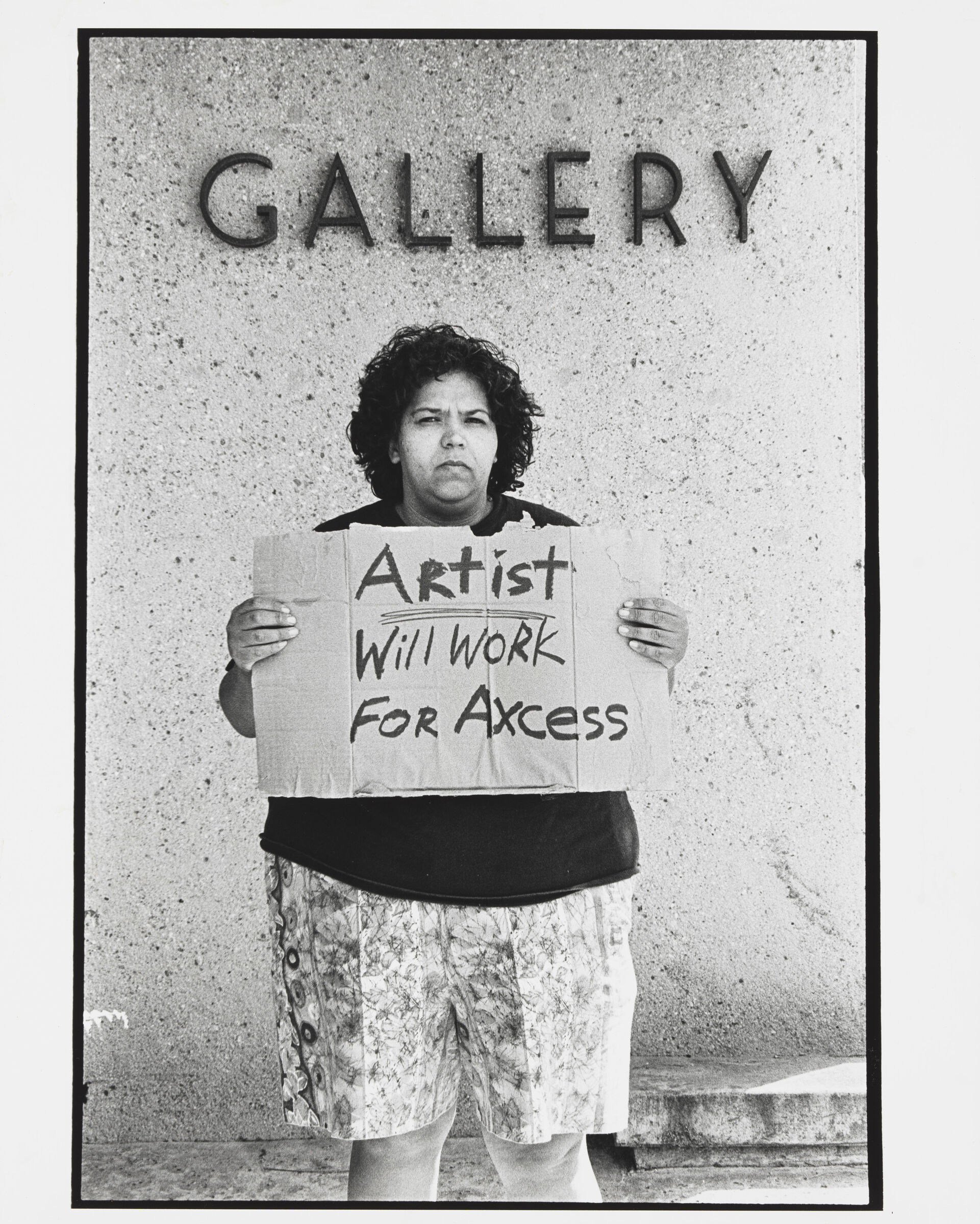Standing in front of sign that says "Gallery," a large Latinx woman holds a cardboard sign that reads "Artist Will Work for Acess." The last word is mispelled.