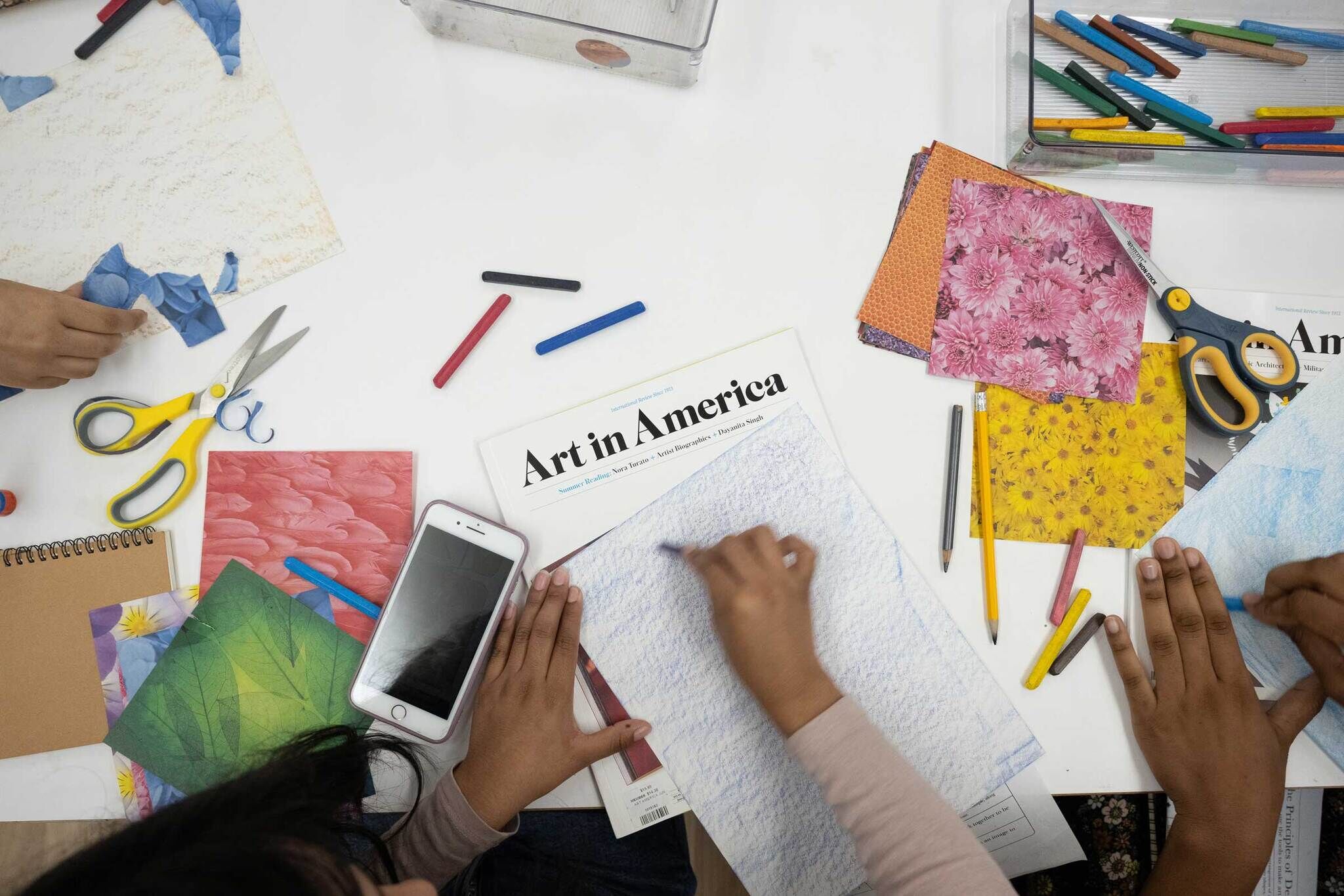 Numerous papers scatted on the paper, one reads 'Art in America.' Arms reach out to cut papers to create a collage.