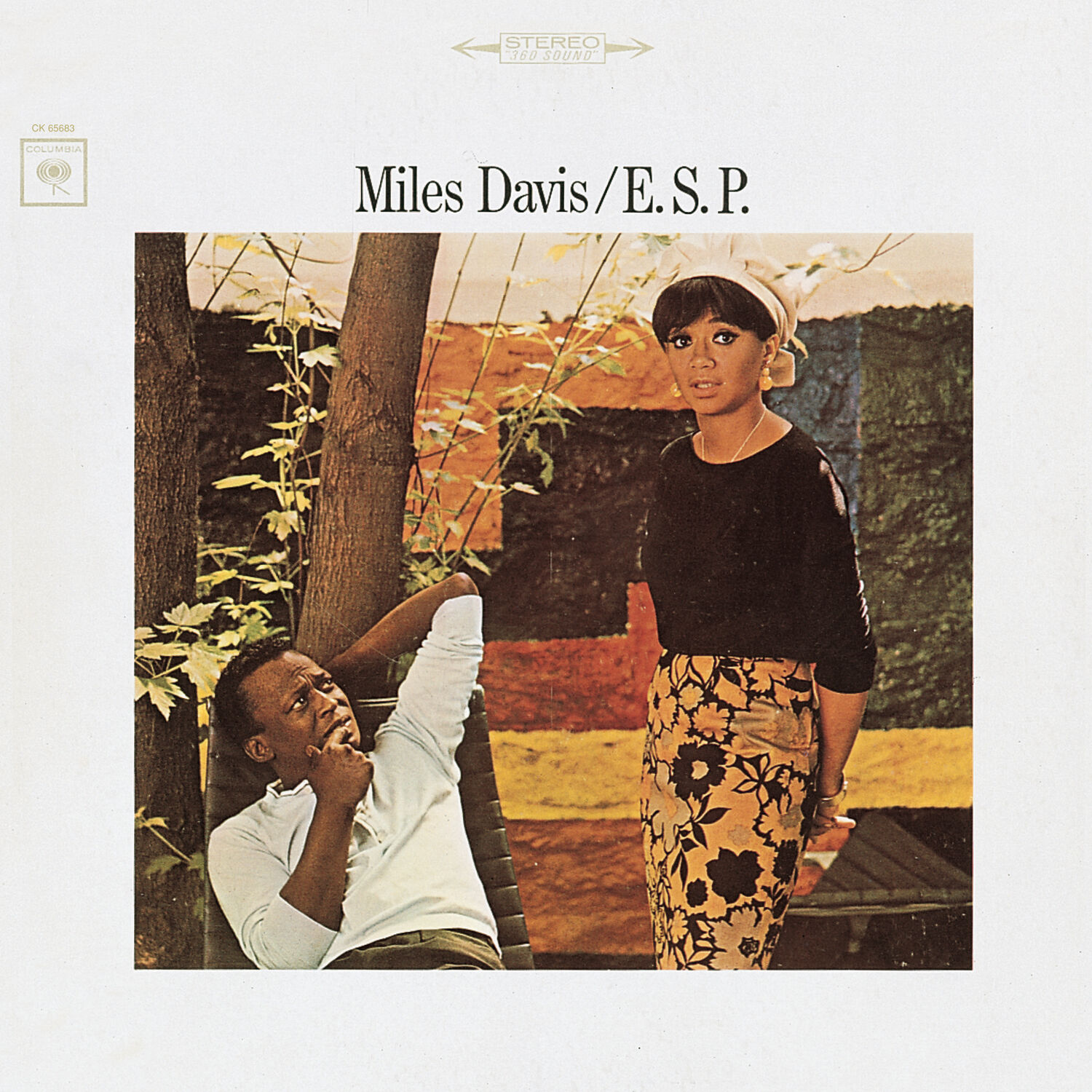 An album cover for Miles Davis' album E.S.P. Miles Davis is laying on a sunchair against a Fall background looking up at his wife in a white hankerchief, black top, and black and yellow skirt with flowers.
