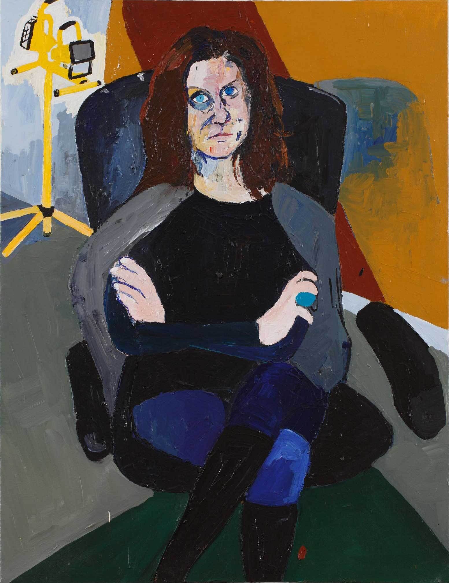 The portrait of a white woman seated with her arms and legs crossed on a rotating desk chair. She has blue eyes, reddish-brown hair, and is wearing a black top, blue jeans, black boots, and a grey jacket draped over her shoulders. She sits in a room with a multicolored wall, grey floors, and a dark green rug. In the background is a tall, yellow work light.