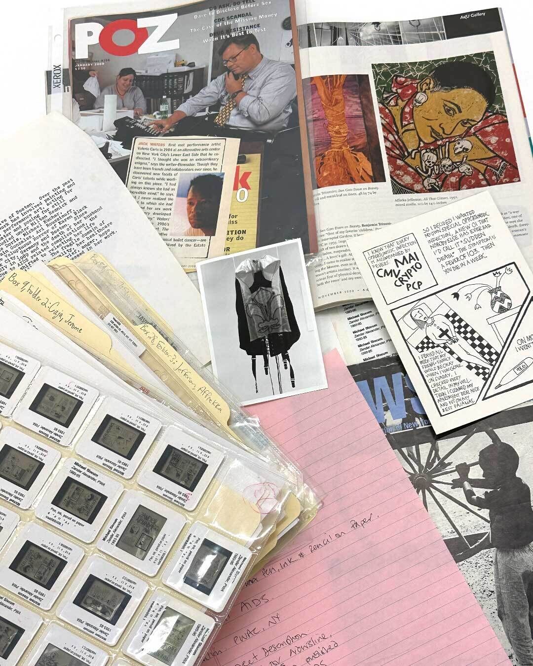 A range of archival materials spread across a white table, including
slides, file folders, typewritten documents, magazine spreads, and a small black-and-white photograph.