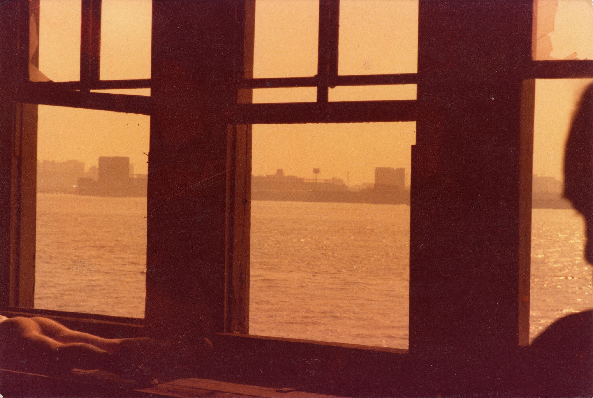 The lower half of a naked person in front of windows overlooking the water all cast in an orange light.