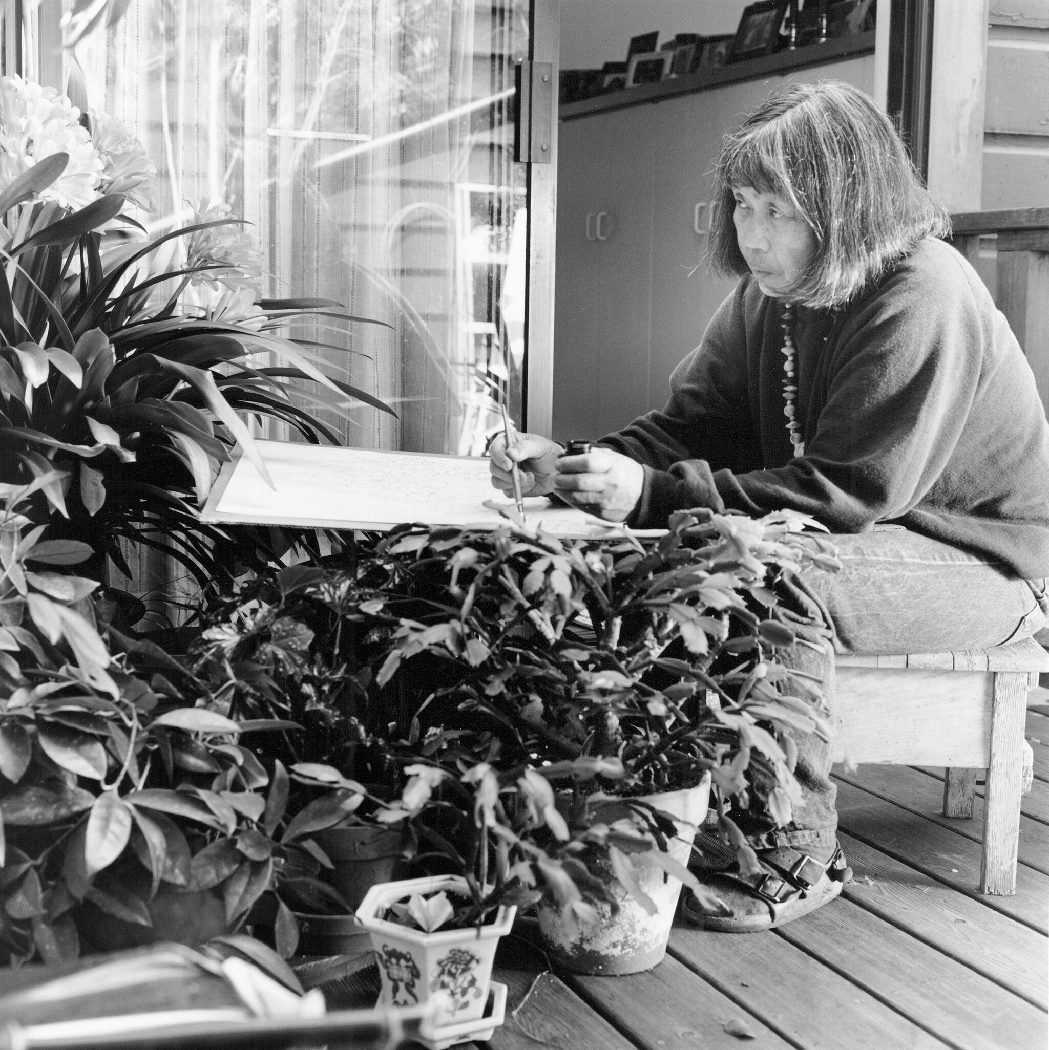 A woman sitting on a wooden deck drawing the plants that surround her. 