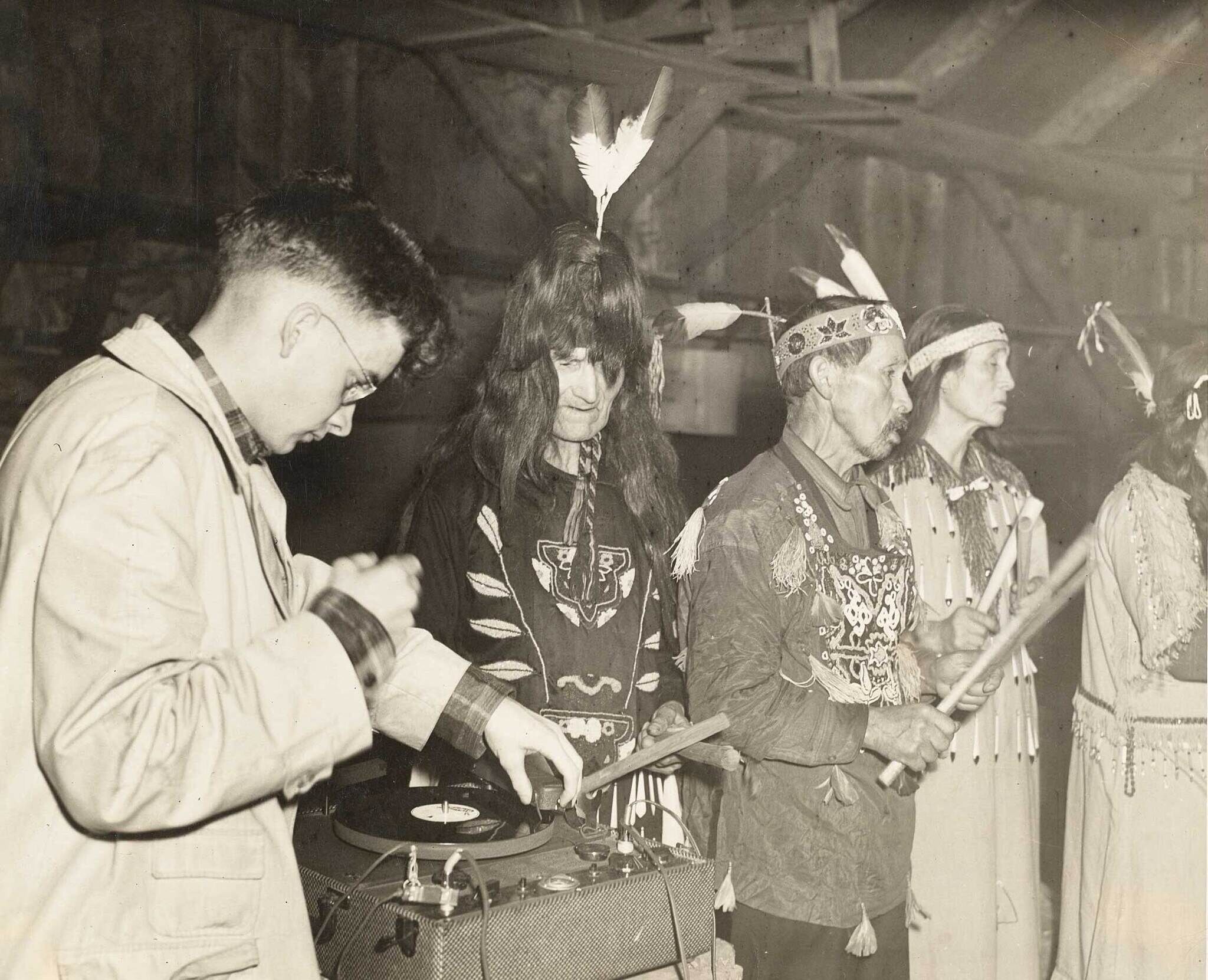 Harry Smith, a young white man with glasses recording four men in traditional Lummi ceremony.
