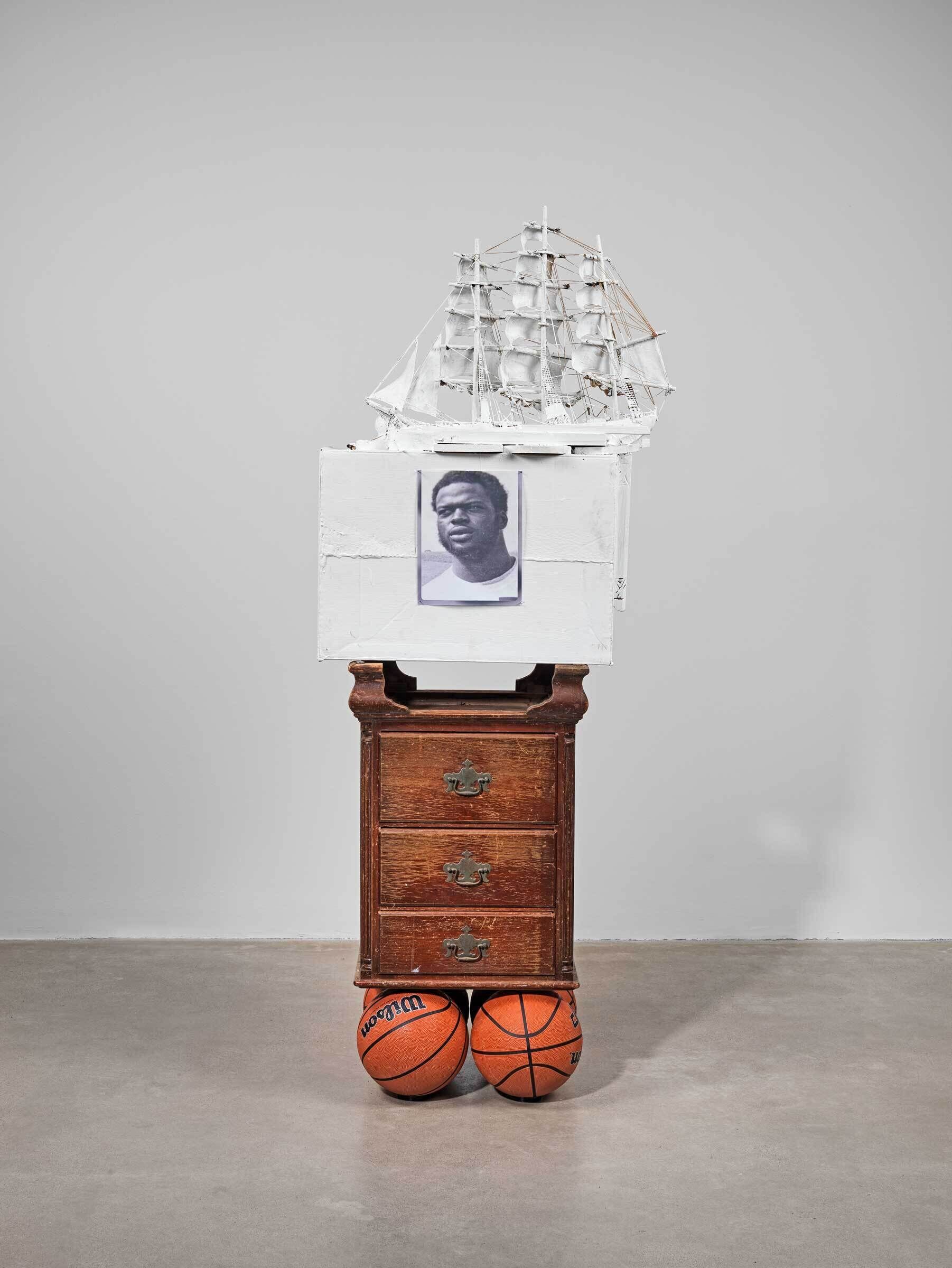 A sculpture consisting of four vertically stacked components, from top to bottom: a miniature ship, a white box with a black and white portrait of a dark skinned masculine figure, a worn wooden dresser, and four basketballs.