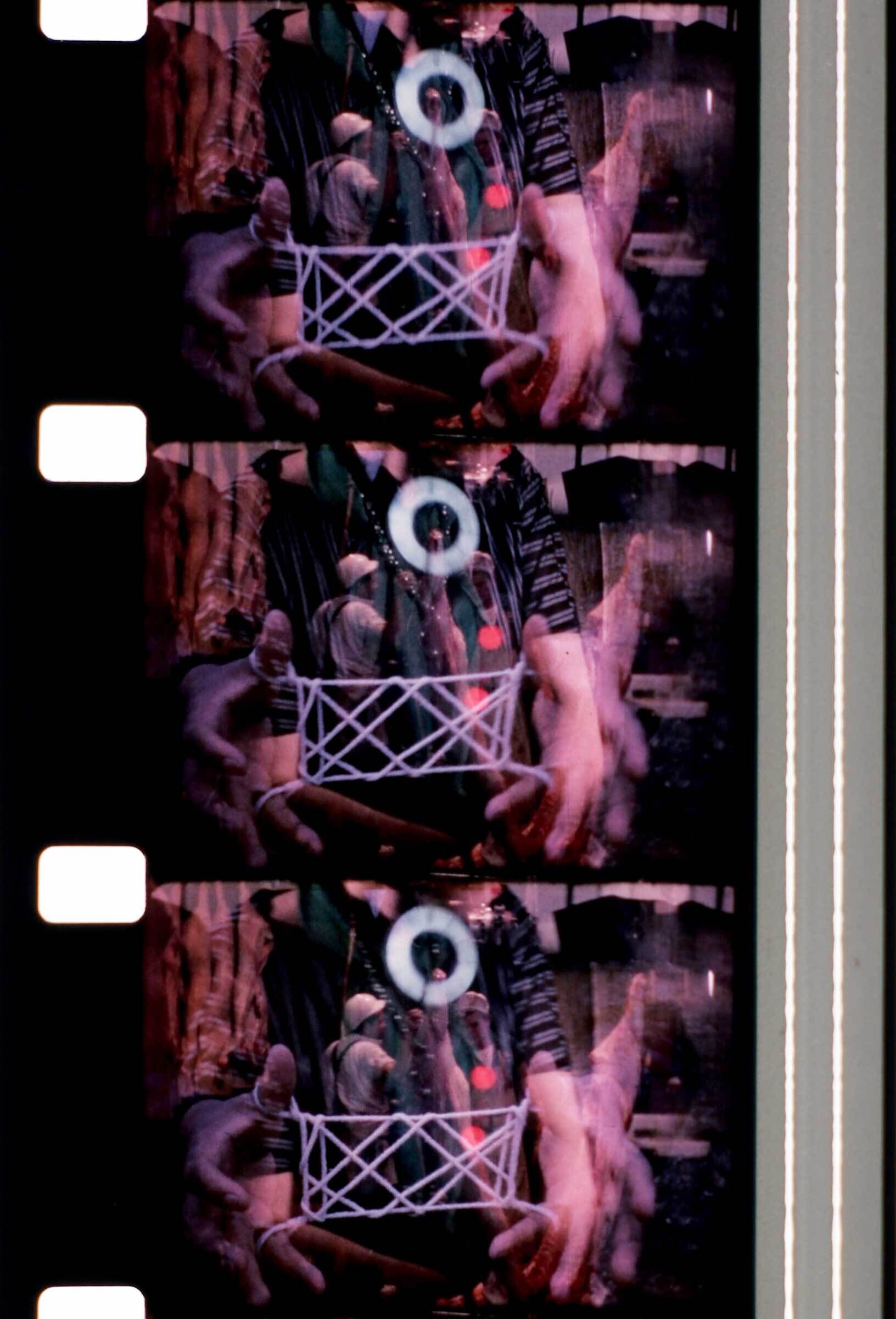 A still image from a film, which has multiple images superimposed on top of each other. Two hands are holding open a string shape.
