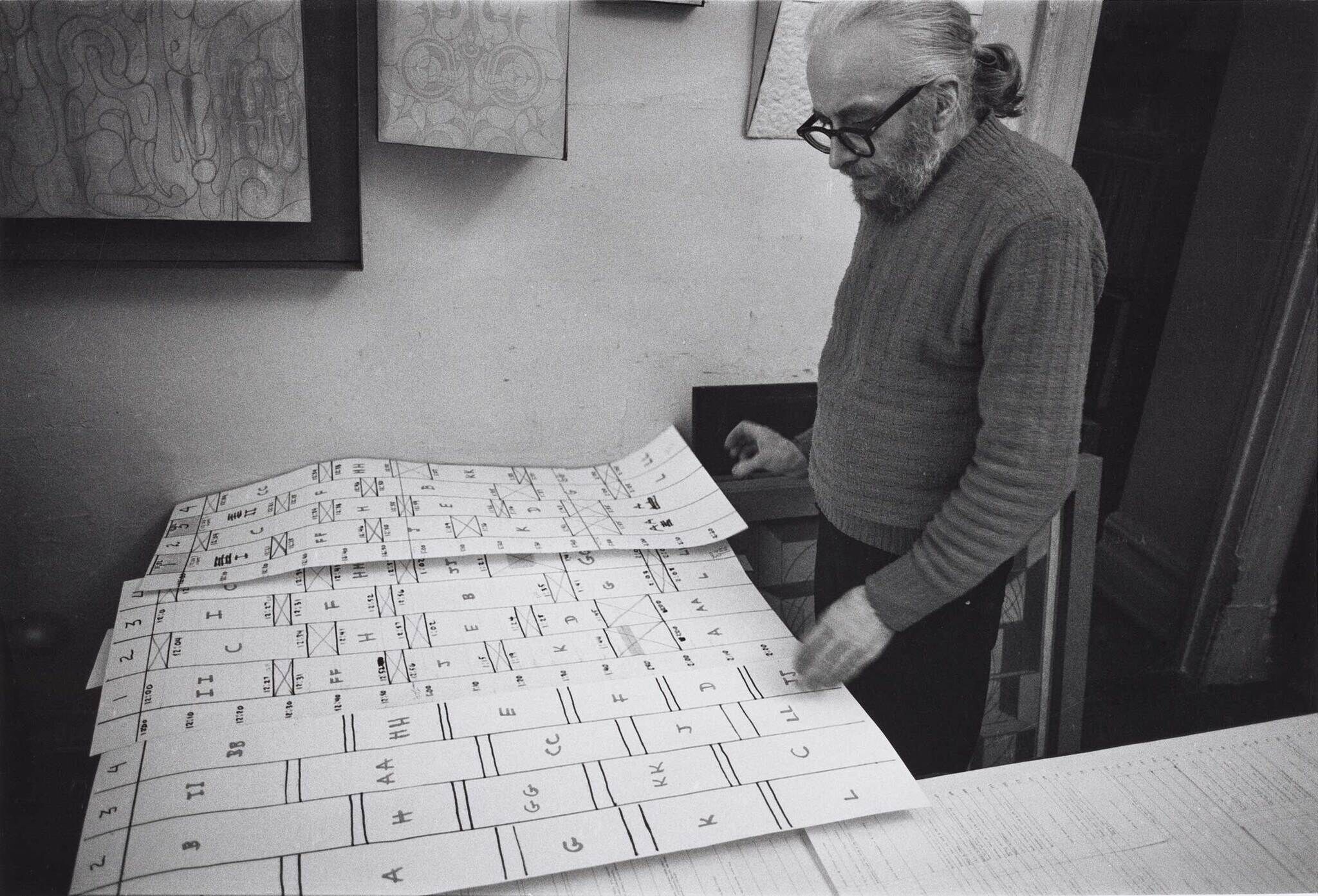 A greyscale photograph of Harry Smith, standing in front of multipe stacked rows of charts arranged in front of him.