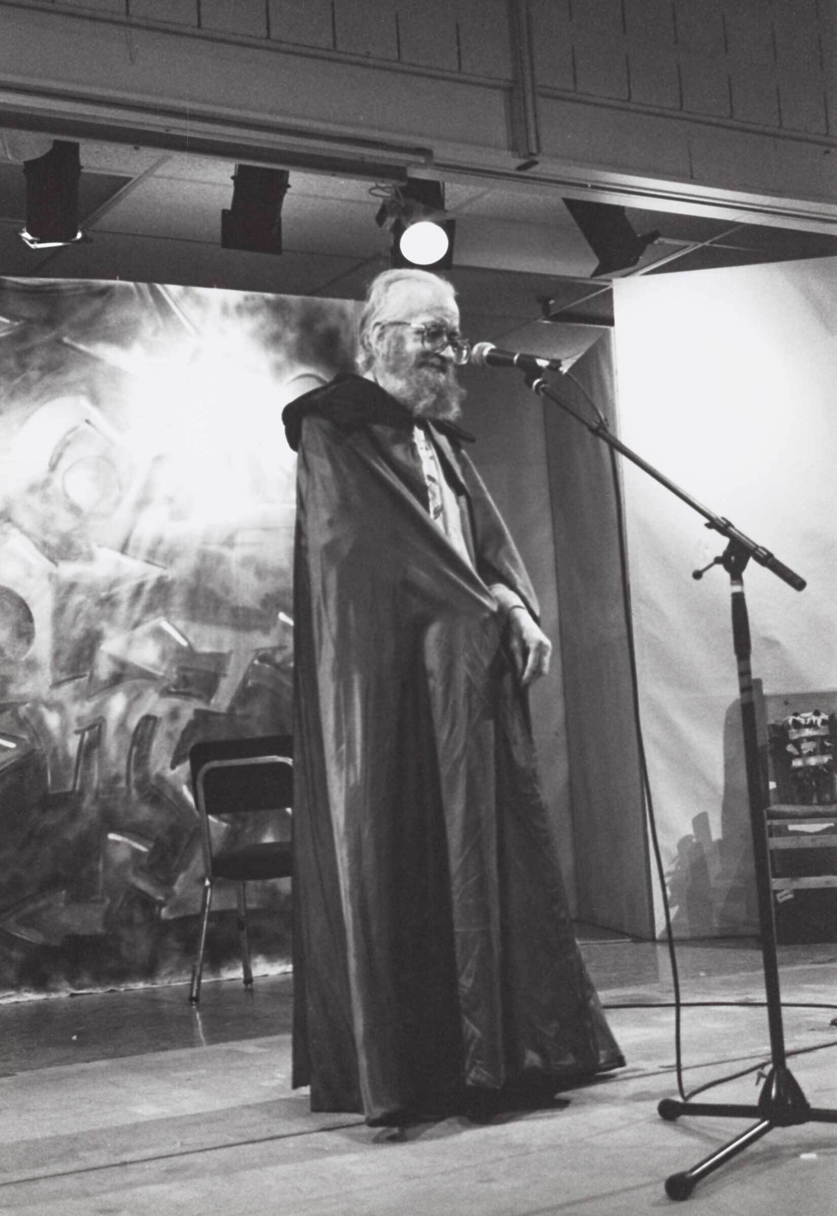 A black and white photograph of Harry Smith with white hair and a long beard. He is wearing round glasses and a long black cape and standing at a microphone in an otherwise empty room. 