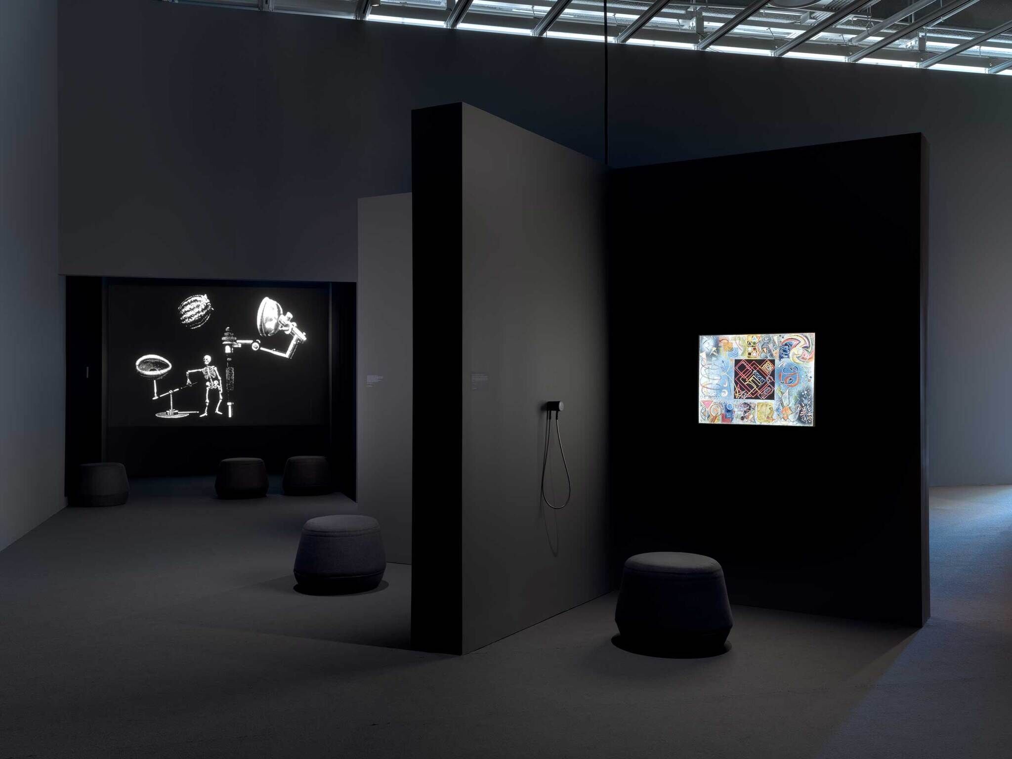 On the left, a dark theater with a skeleton playing on the screen, and on the right is a pinwheel-like structure with a lightbox illuminating a colorful painting and speaker on its left.