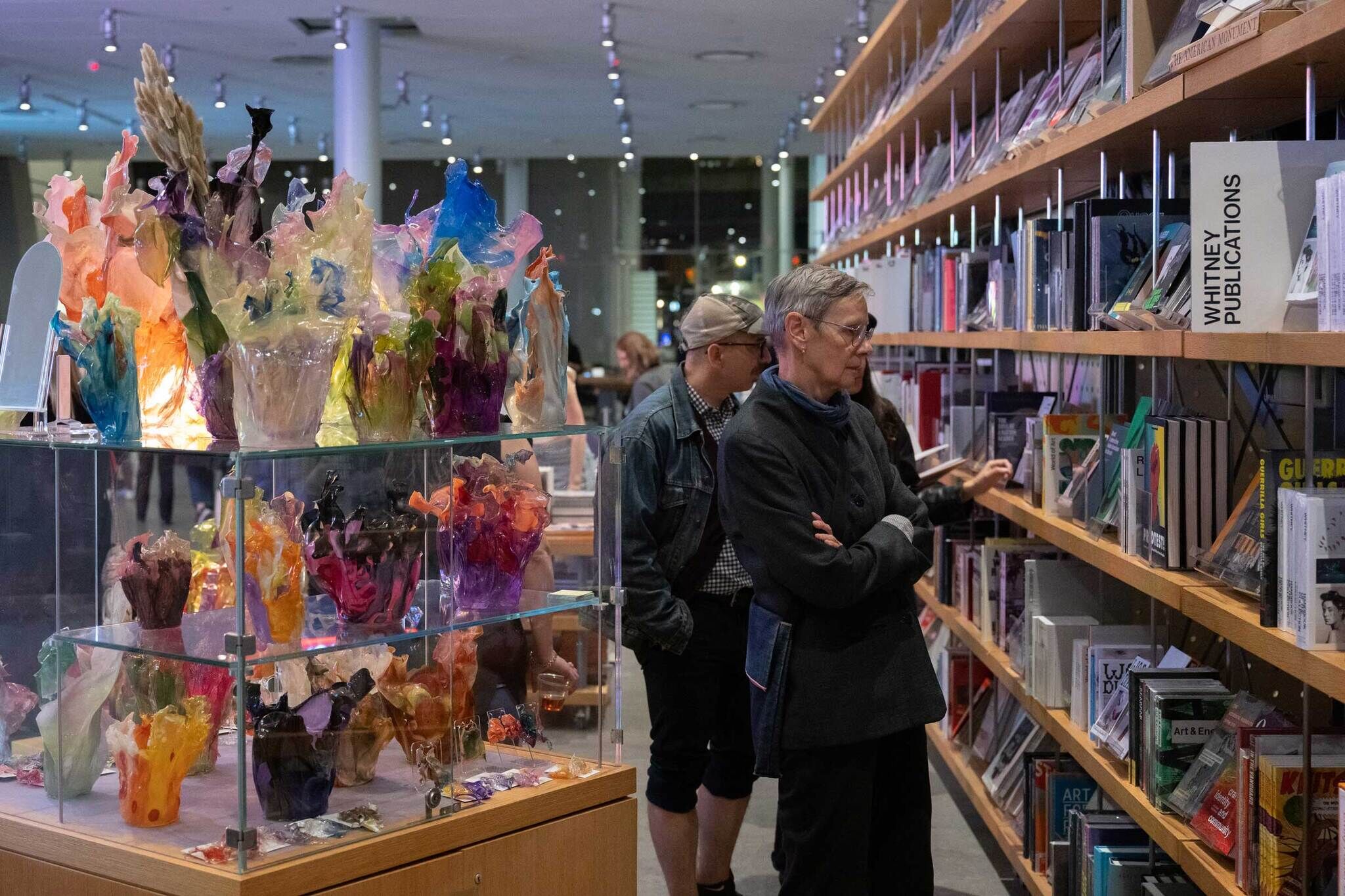People stand looking at bookshelves in the Whitney Shop. Next to them, is a display of colorful glass vases on clear shelves.