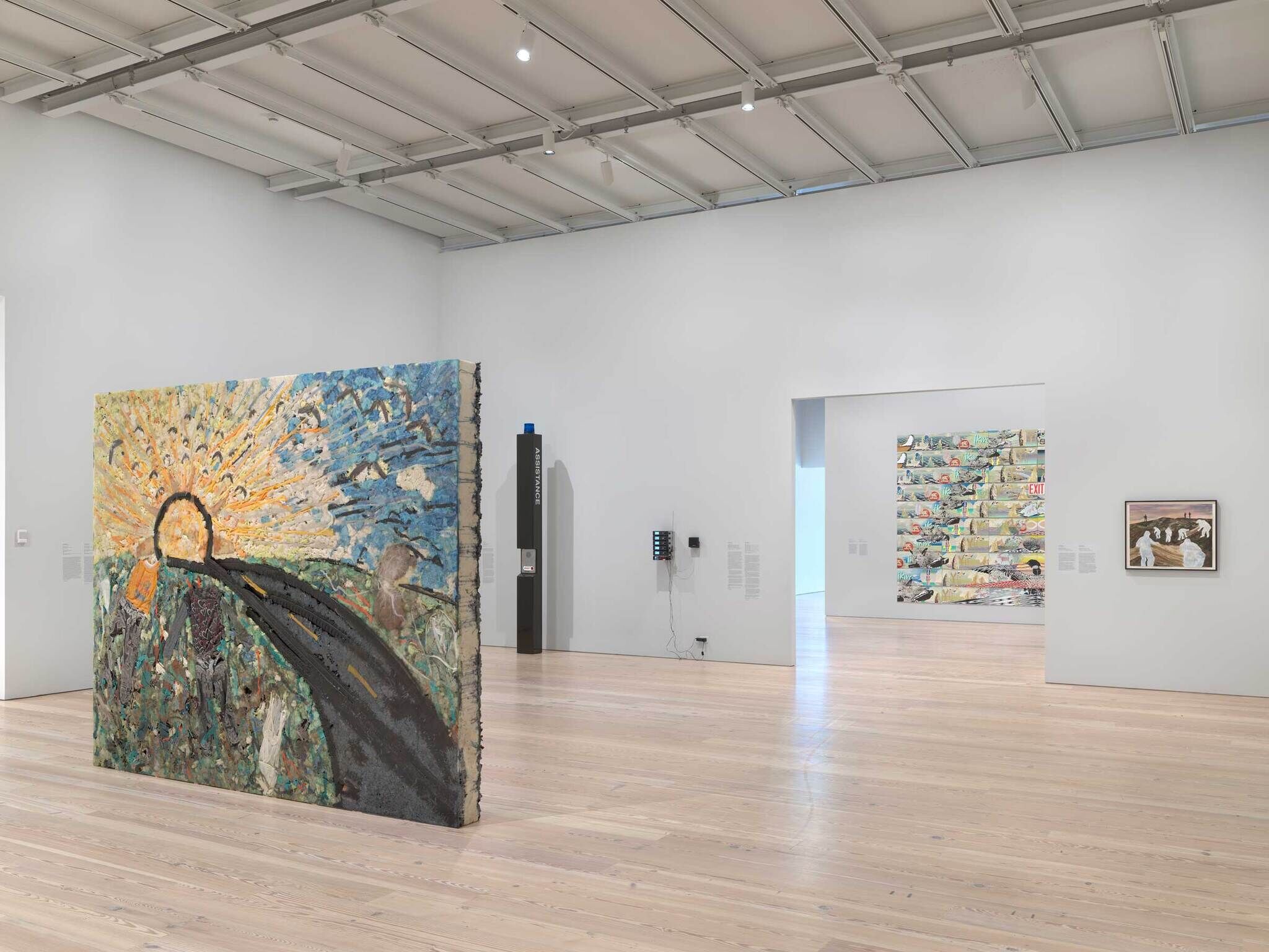 A big painting of a sun and a road standing in the middle of the room. In the background there is a emergency blue light pole and through the open doorway into the other gallery there are more paintings.