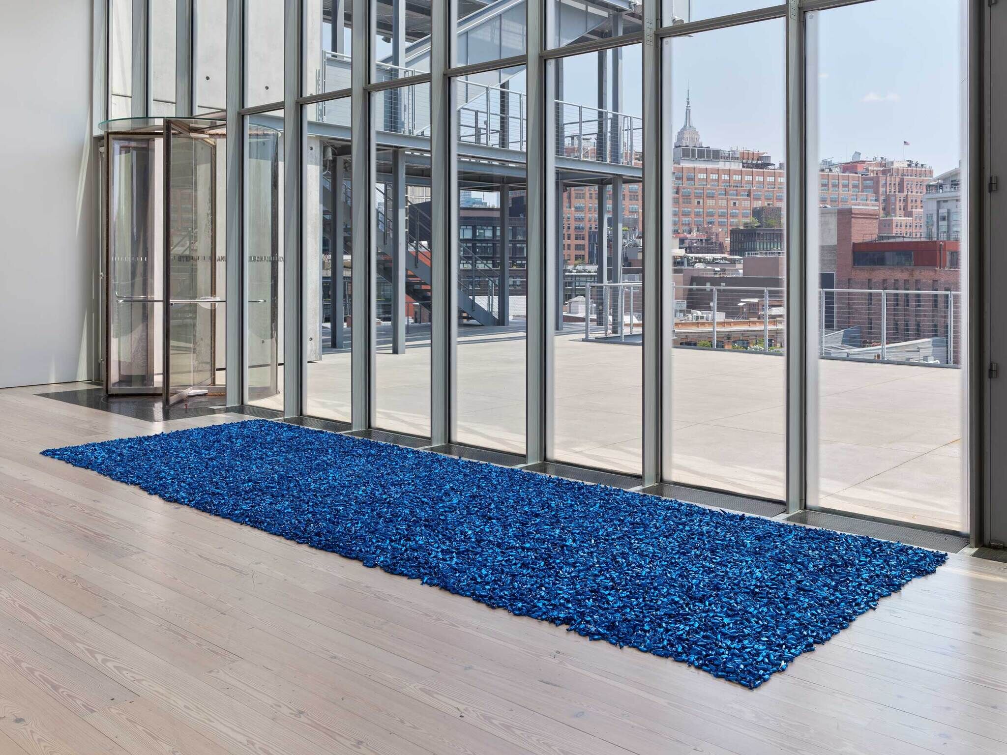 A huge pile of small blue wrapped candy on the ground in front of big floor to ceiling windows showing a balcony and the New York skyline.