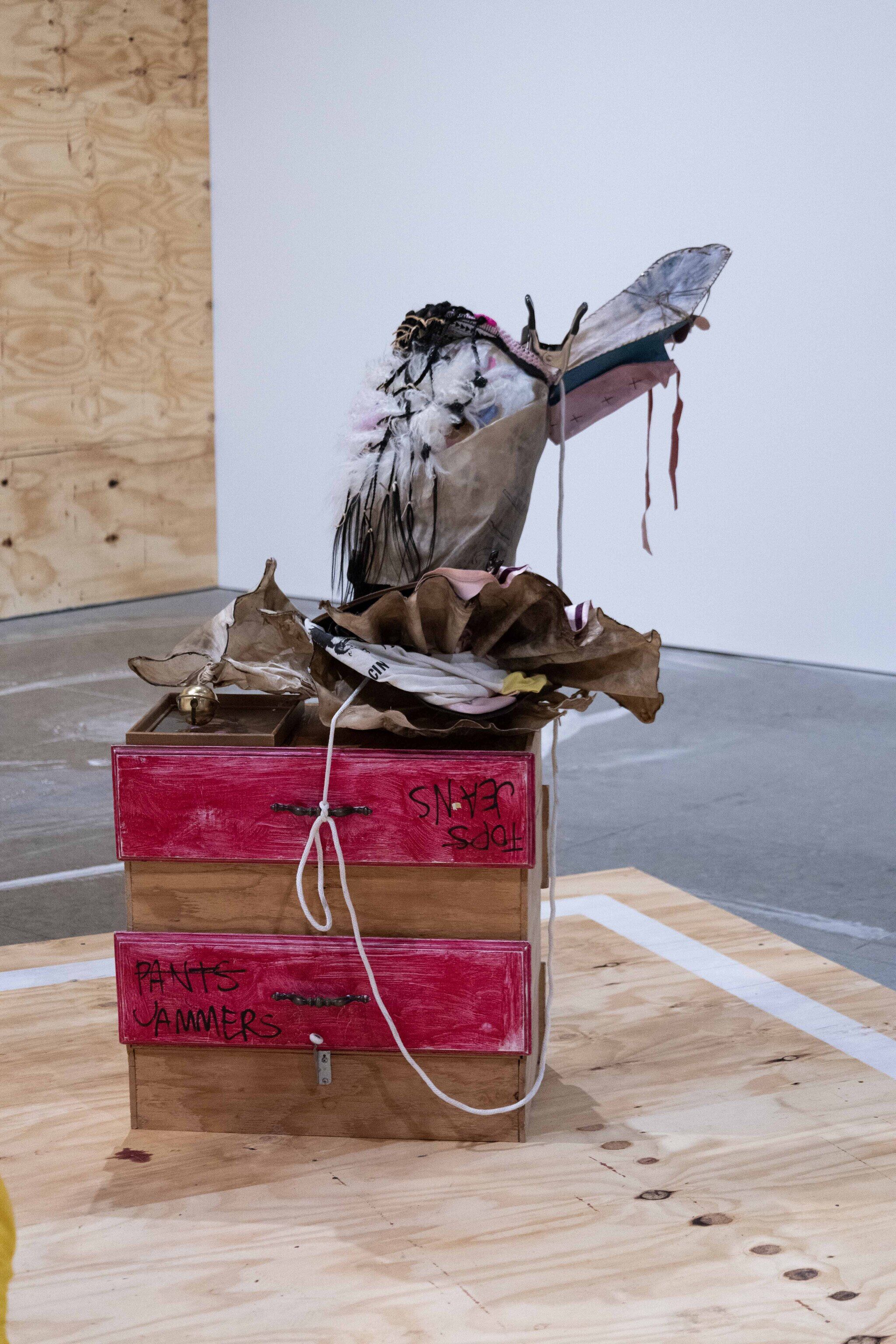 This work features boxes stacked with elk rawhide, wood, cotton, satin, artificial hair, sheep hair, framed poster, metal, tattoo ink, pastel, makeup, oil, leather, metal clamps, yarn, plastic beads, glass beads, felt, plastic, rope, and graphite leaning asymmetrically on the top. 