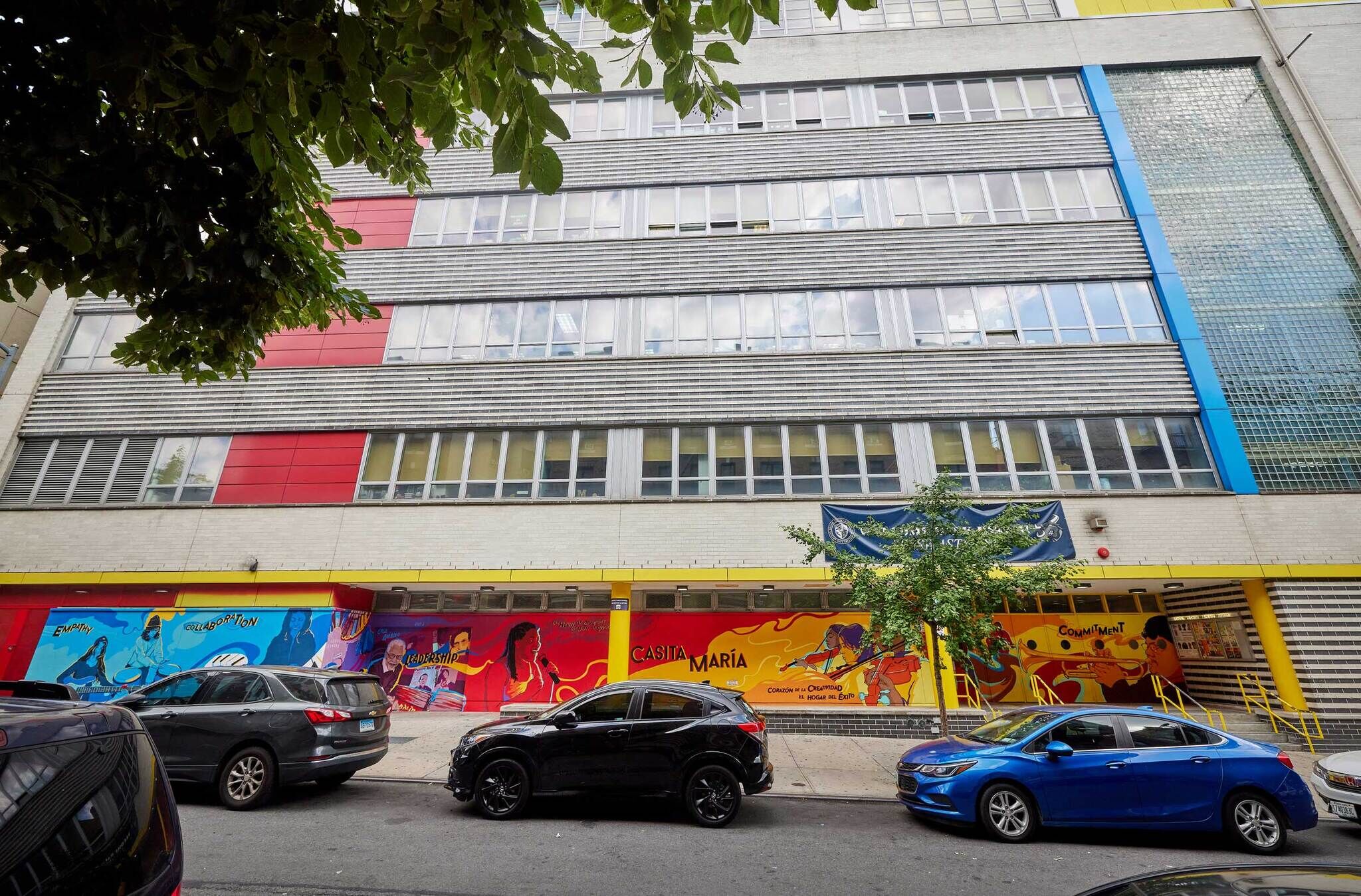 A building with colorful bright murals on the side, cars parked in the road in front of it.