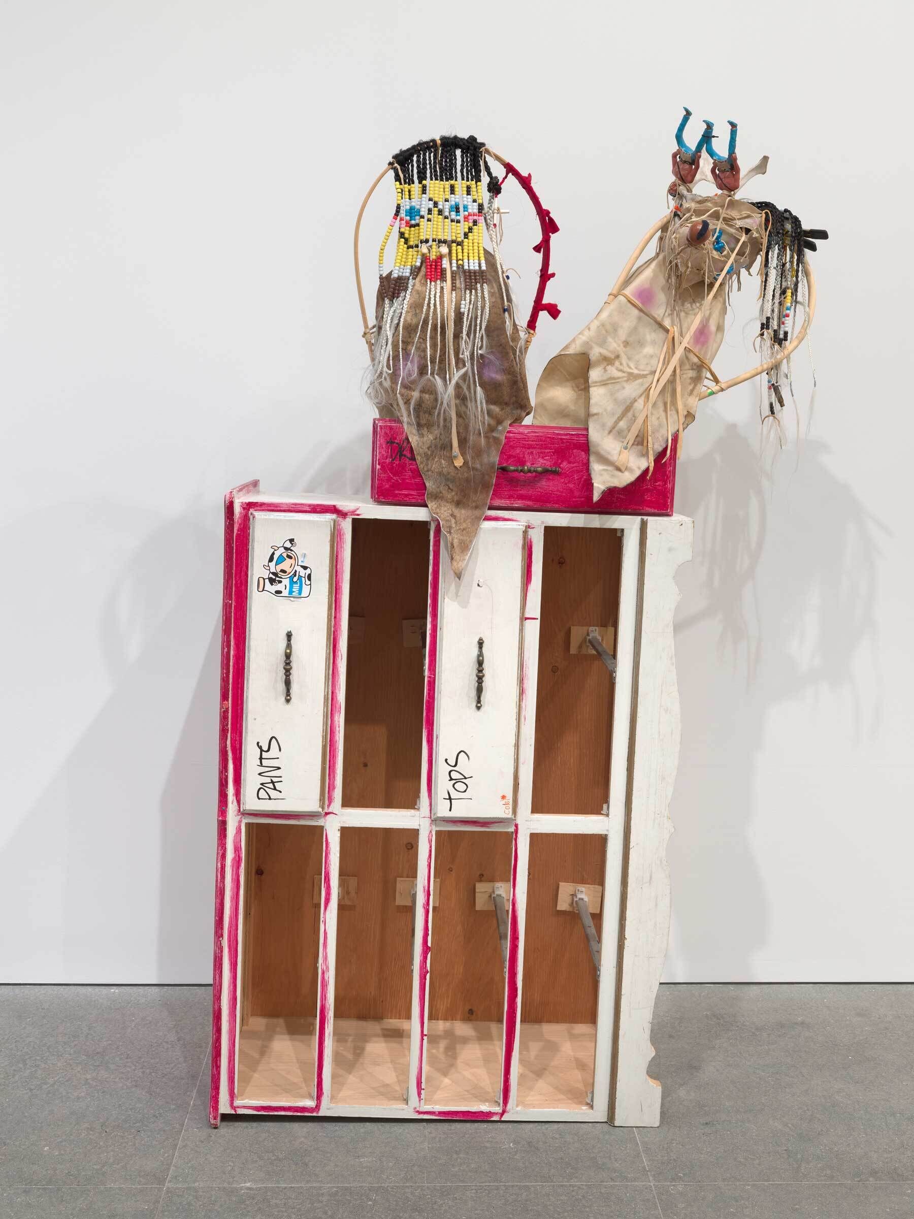 An art installation featuring a wooden wardrobe with colorful, abstracted figures on top, labeled "PANTS" and "TOPS."