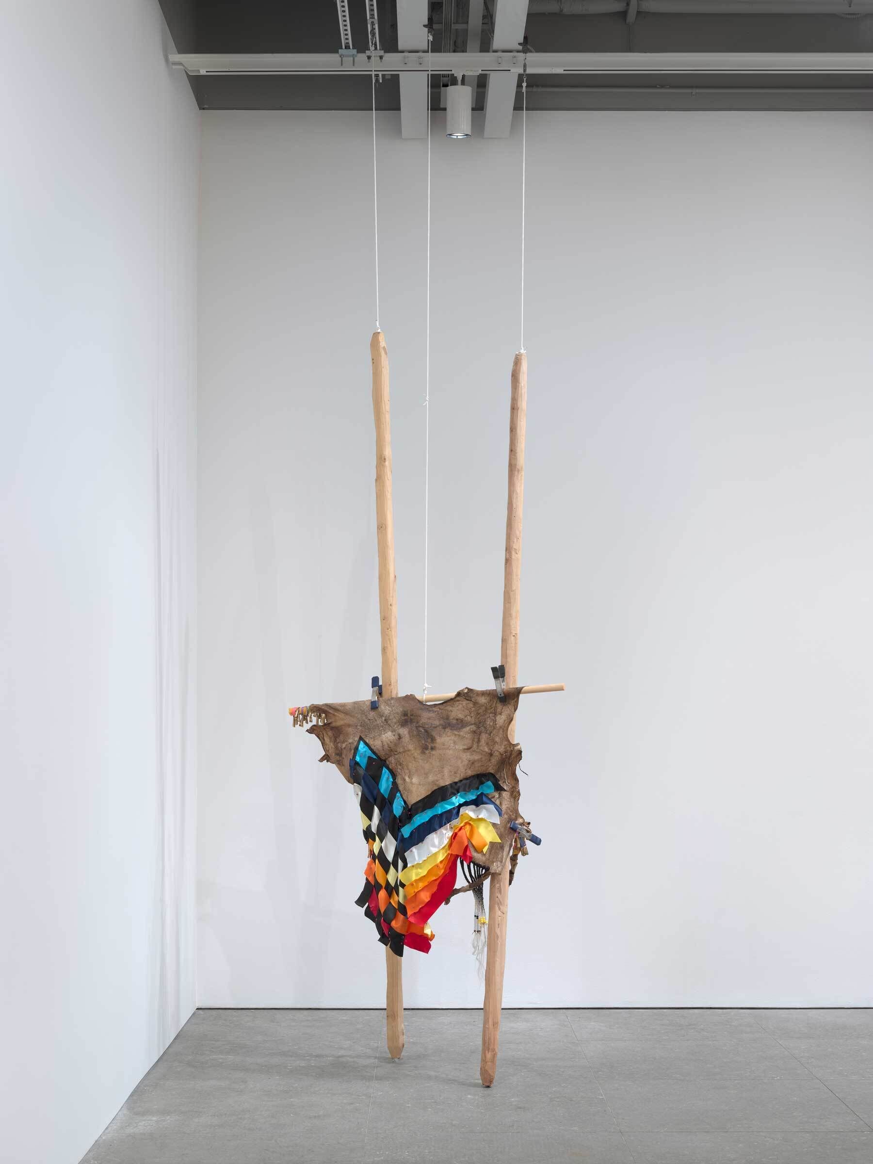 Modern art installation with colorful fabrics hanging from a wooden structure suspended by ropes in a white gallery space.