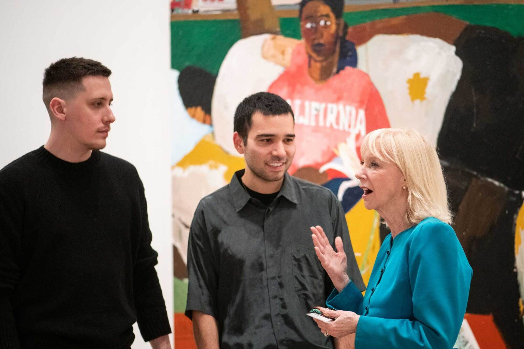 Image of a gallery in Henry Taylor: B Side with a woman in blue speaking to two men in gray and black in front of an artwork featuring two subjects dressed in casual clothing on a couch.