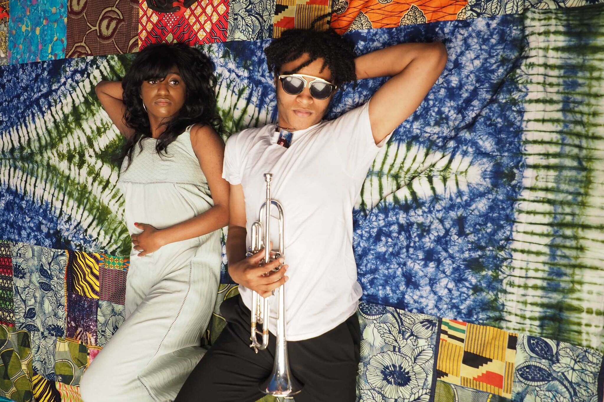 A Black man and woman lay on a multicolored tie-dyed quilted blanket. The man is wearing sunglasses and holds a trumpet.