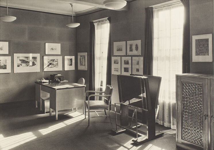 Vintage office with sunlight casting shadows, a desk, chair, artworks on walls, and a large open window.
