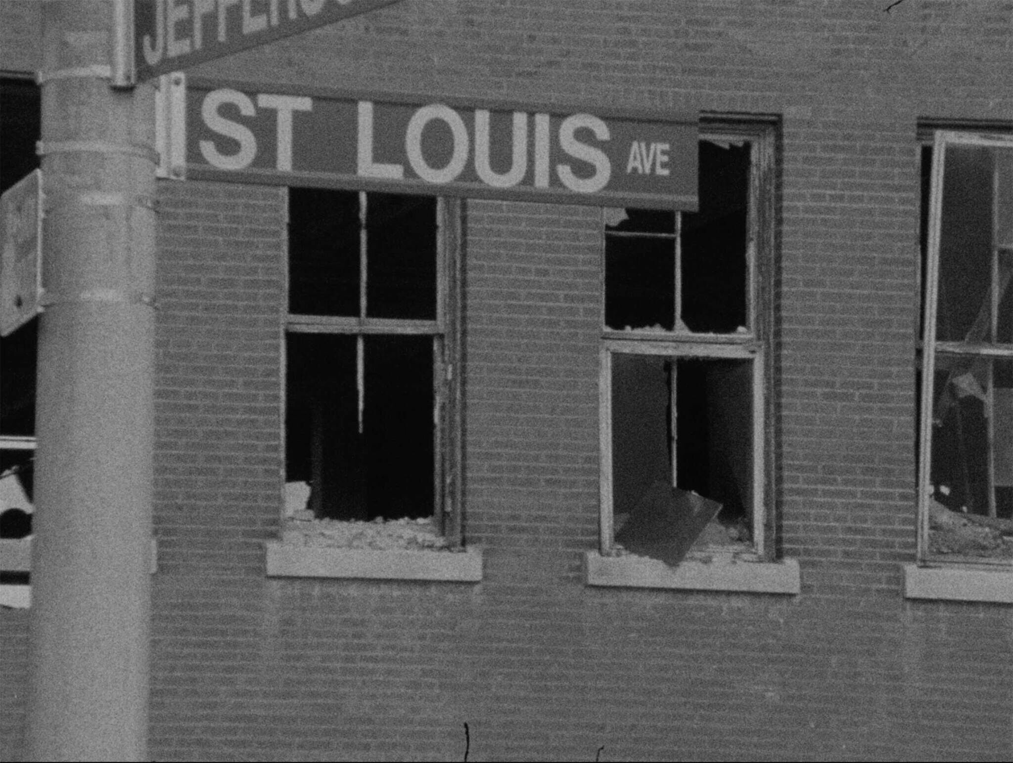 Black and white photo of a building corner with "1ST LOUIS AVE" sign, broken windows, and visible disrepair.