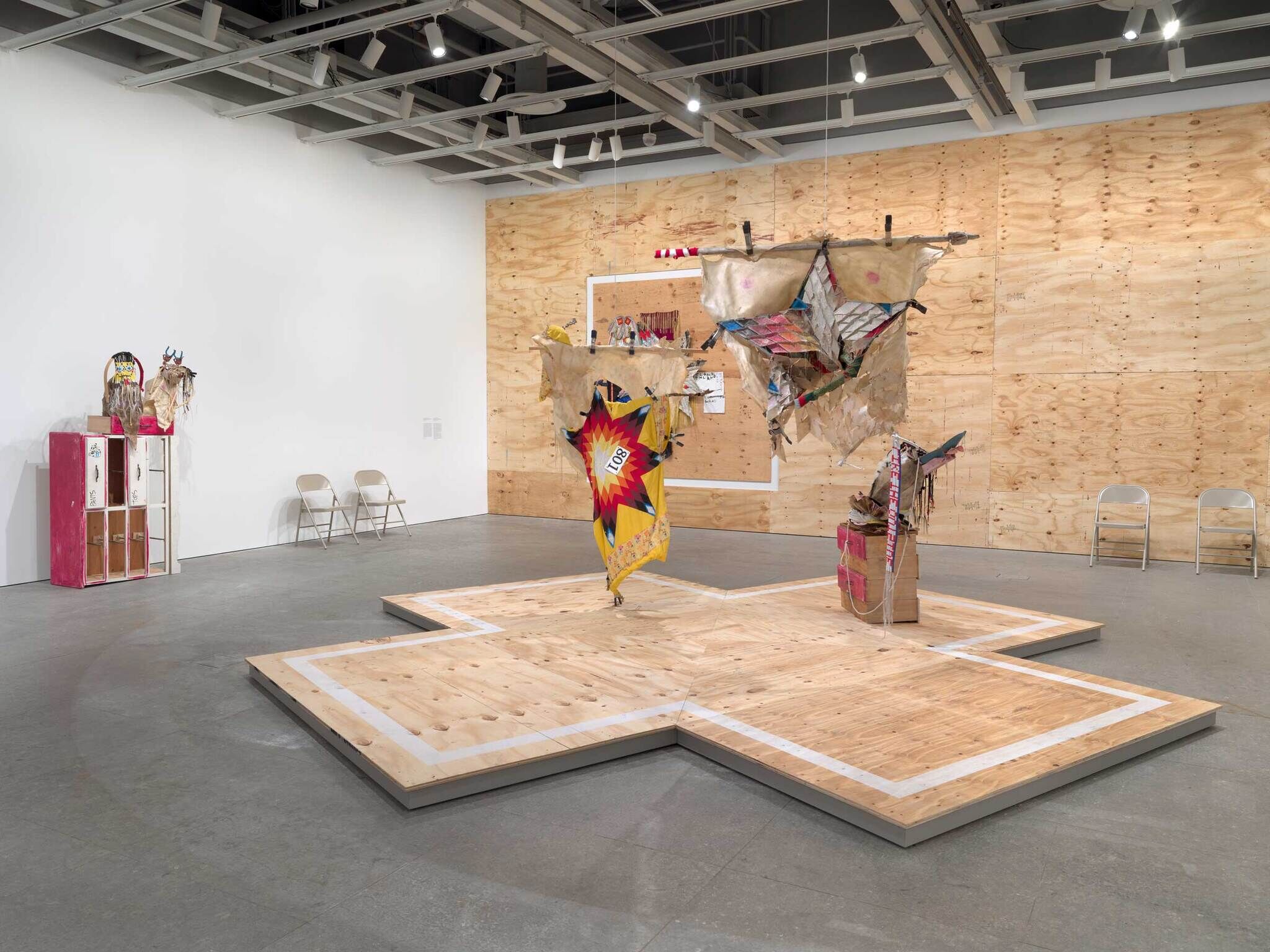 Several colorful artworks in materials including fabric and wood set on a platform and against a backdrop of plywood. 