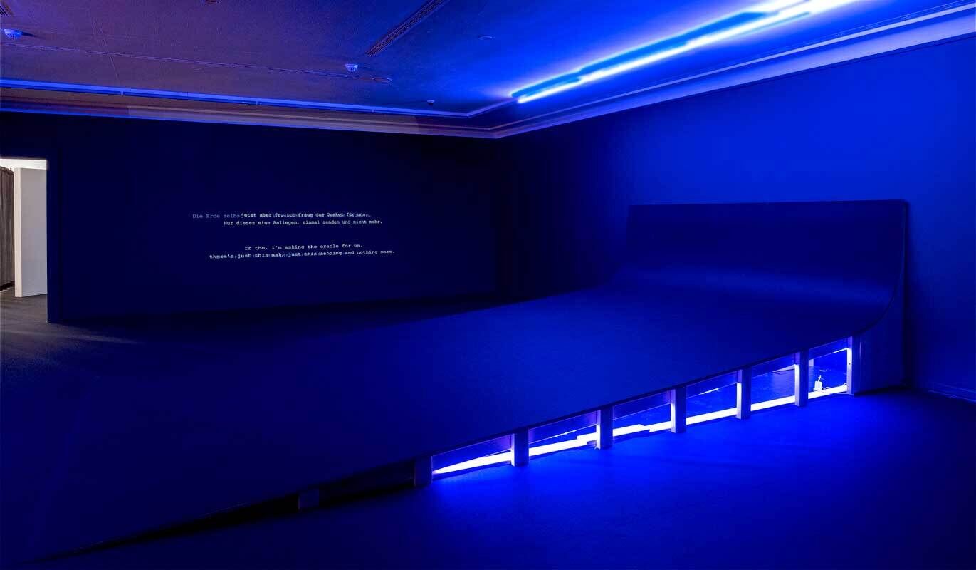 A dimly lit room with blue lighting, featuring text projected on a wall and a curved bench-like structure.