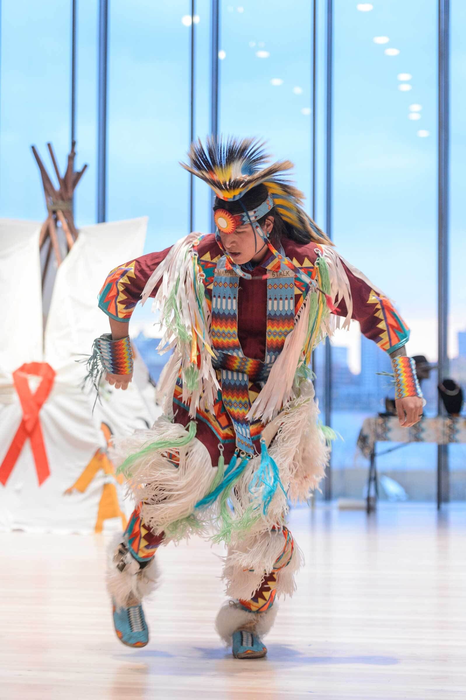 Indigenous dancer in traditional regalia performing at an indoor event, with a tipi in the background.