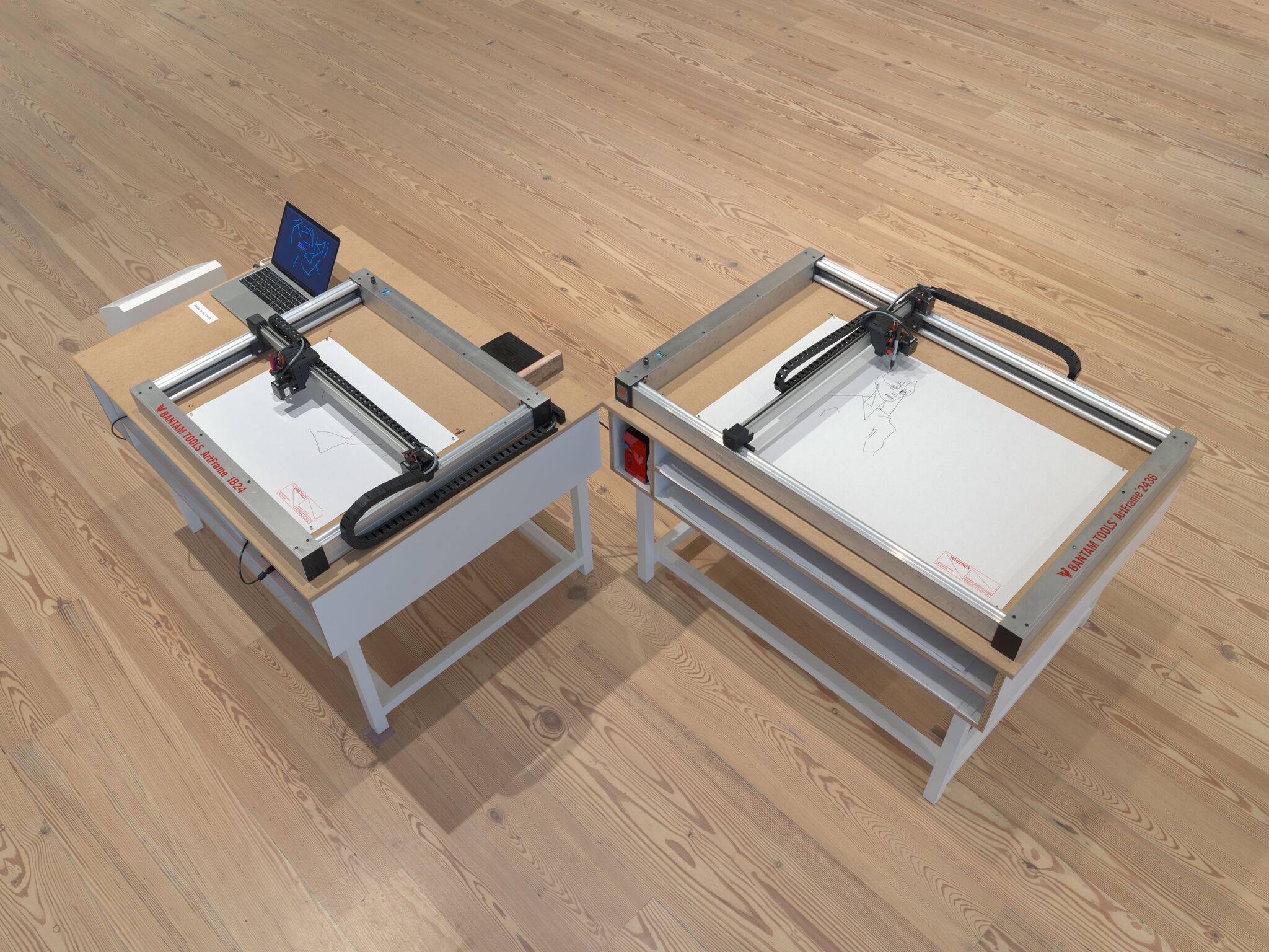 Two robotic drawing machines sketching on paper atop tables in a room with wooden flooring.