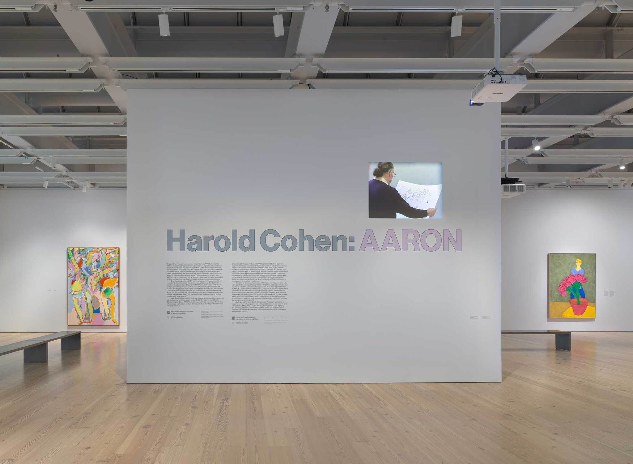 Exhibition space with "Harold Cohen: AARON" display, colorful abstract paintings, and a video projection of an artist at work.