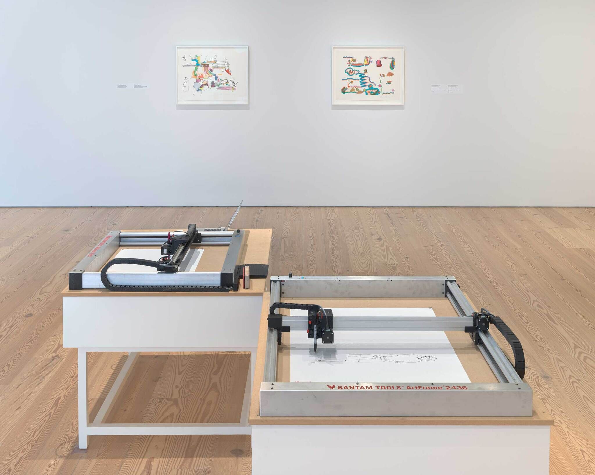 Active plotters drawing line images with black marker on display in an art gallery with framed artworks on the wall.