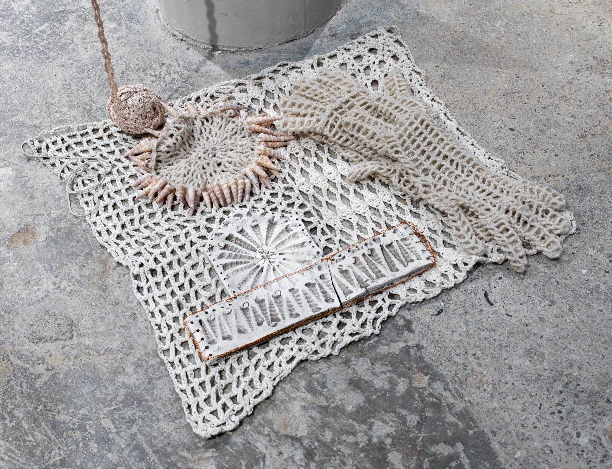 A crocheted textile artwork resembling a human figure with a stone, netted gloves, on a concrete floor.