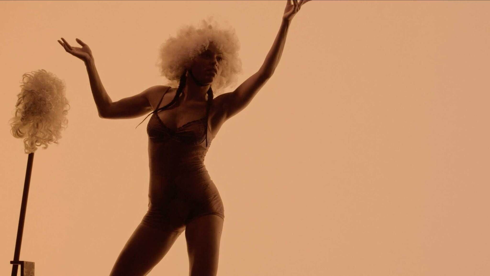 Silhouette of a person with an afro dancing next to a wig on a stand, all cast in a warm, orange hue.