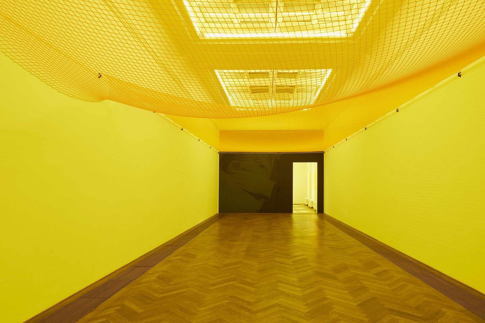 A room with yellow walls and ceiling, a net hanging below white lights, and a herringbone wood floor leading to an open door.