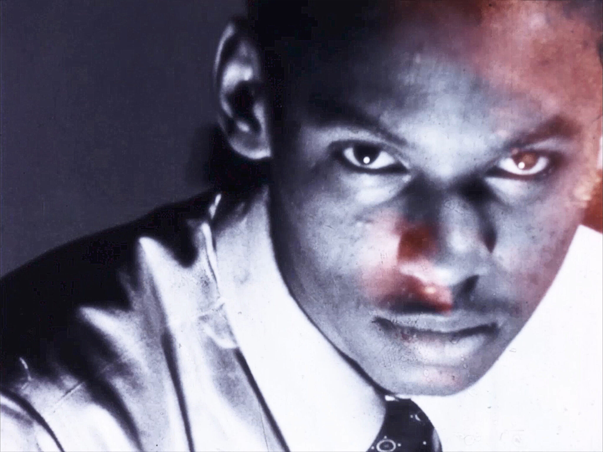 Close-up of a man with intense gaze, wearing a shirt and tie, with a shadowy, reddish overlay.